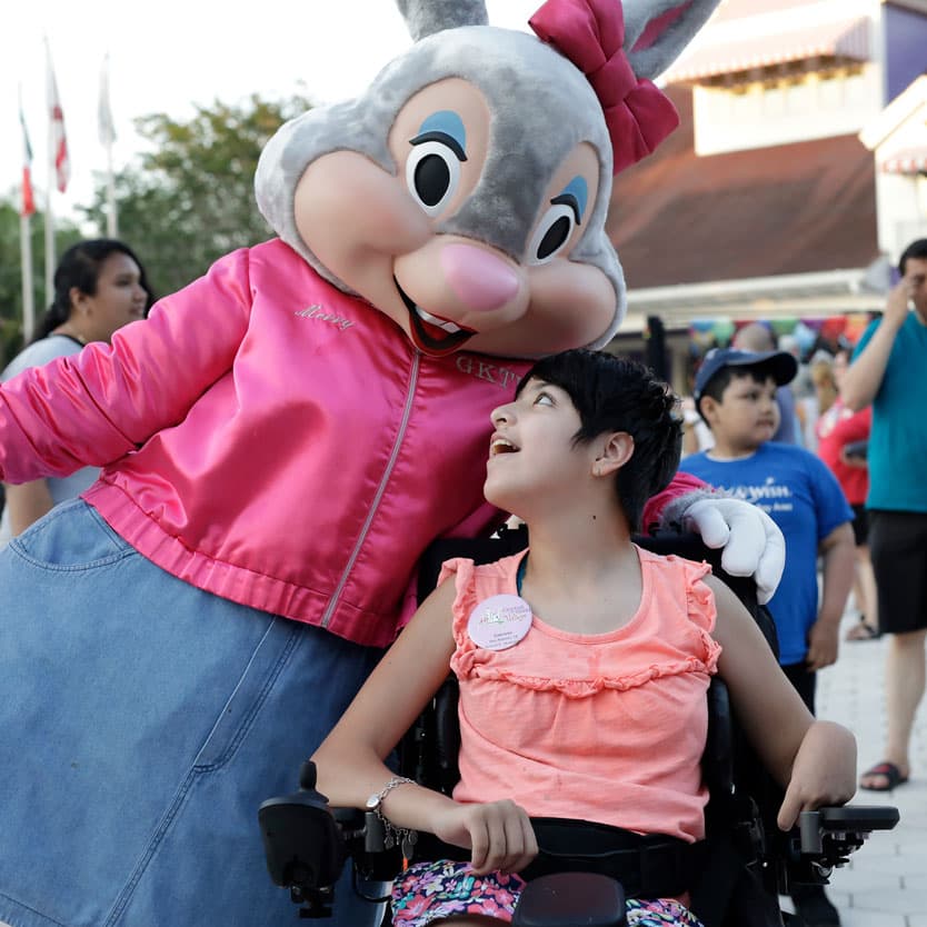 A costumed rabbit character with a pink bow, in a pink satin jacket embroidered Merry and lettered GKTW, leans down beside a young girl seated in a black wheelchair, who is looking up at the character and smiling, with other guests and buildings behind them.
