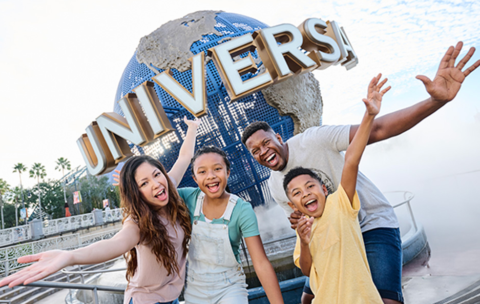 A family of four throws their arms up and laughs in front of the large Universal globe on its mist-shrouded base at the park entrance, with palm trees and a bright sky behind them.