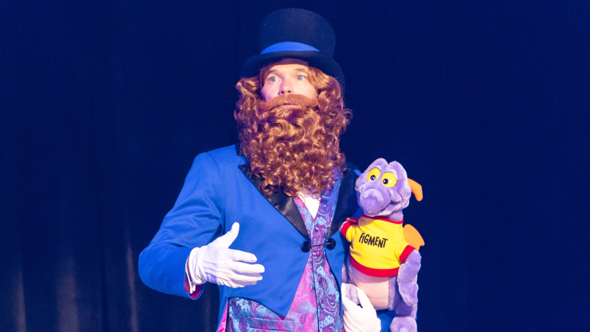 A performer in full Dreamfinder costume, a royal blue tailcoat with black lapels, a pink and teal paisley waistcoat, white gloves, a long curly auburn wig and beard and a dark navy top hat with a bright blue band, holding a plush Figment in a yellow shirt lettered FIGMENT, against a dark navy stage curtain.