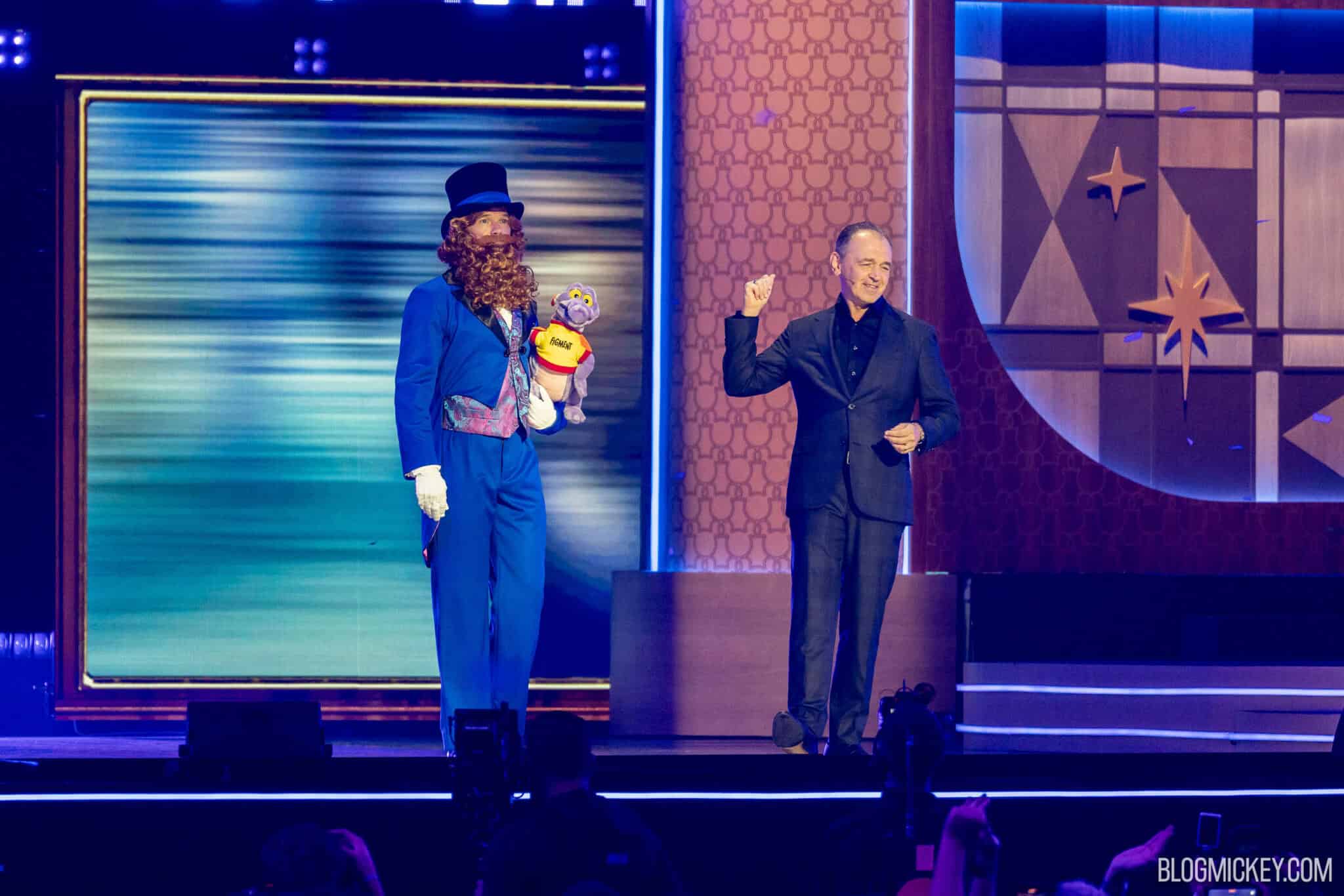 Two men on a brightly lit stage: at left the performer in the blue Dreamfinder costume and top hat holding the Figment plush, at right a man in a black suit and open collared black shirt wearing a headset microphone, raising a fist and smiling, with blue confetti falling and audience silhouettes along the bottom edge.