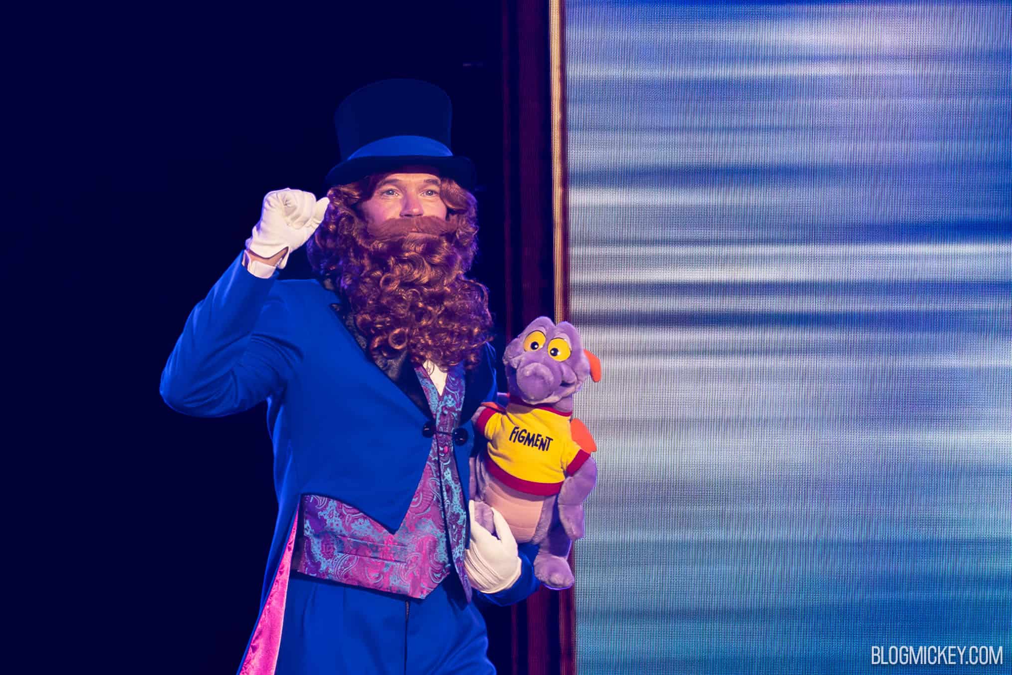 The same costumed performer with a white gloved fist raised beside the brim of his blue top hat, the plush Figment held at his side in his other hand, standing in front of a large blue and white stage screen.