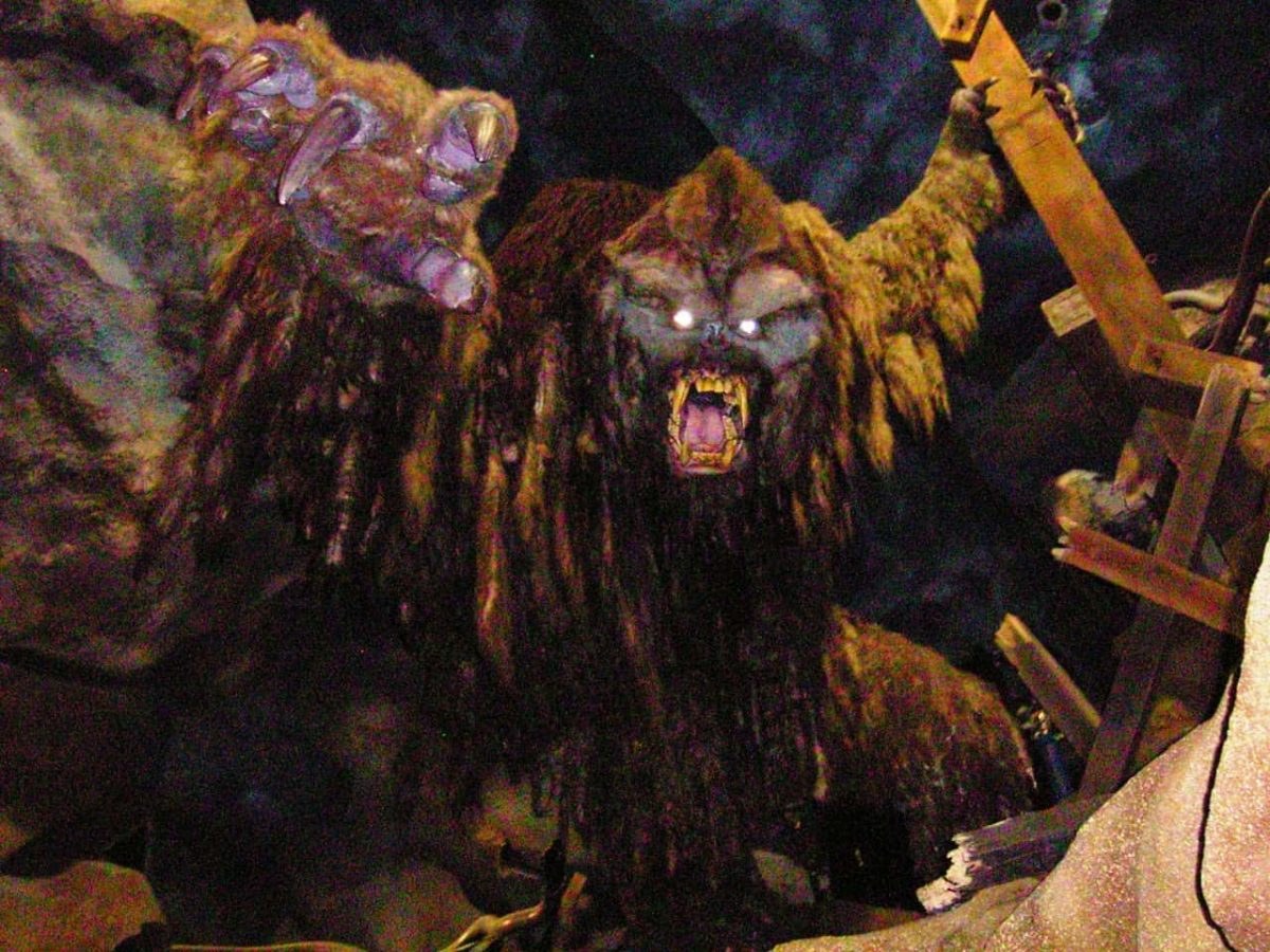 The Expedition Everest Yeti figure in dim amber light, mouth open in a roar with glowing white eyes, both arms raised, one of them reaching toward a broken wooden track structure