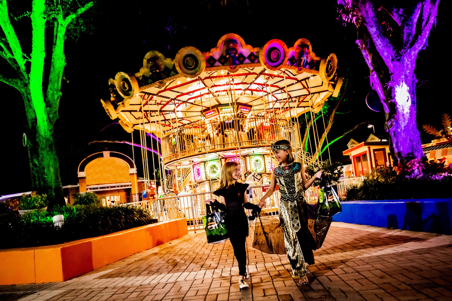 Two children in Halloween costumes carrying treat bags walk hand in hand past a brightly illuminated double decker carousel at night, with park trees uplit in green and purple behind them.