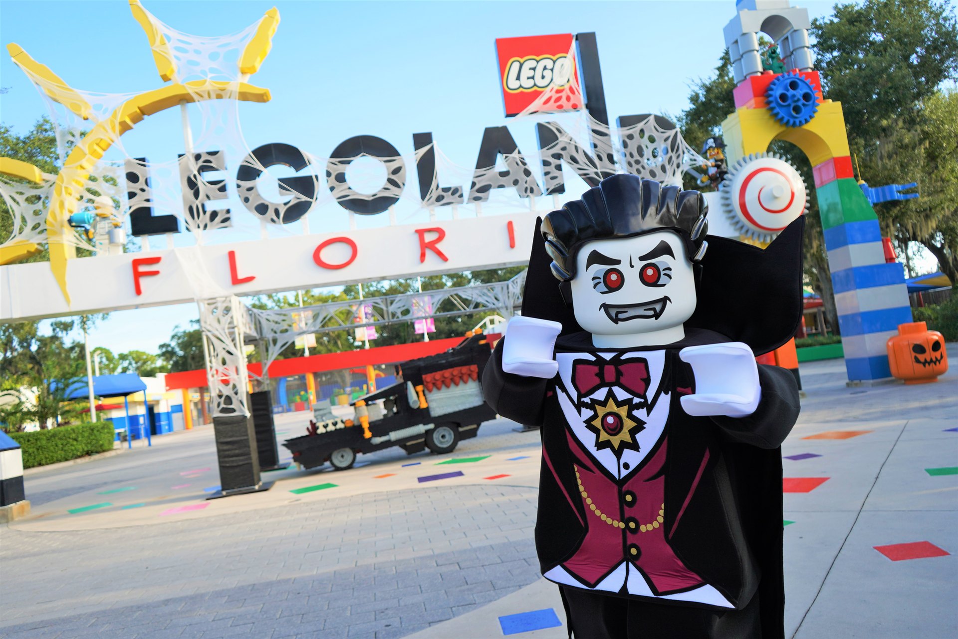 A costumed Lord Vampyre character in a black cape and dark red waistcoat poses in front of the LEGOLAND FLORIDA entrance sign, which is draped in white spiderweb netting, with a large orange Jack-O-Lantern LEGO head standing on the pavement to the right and a brick built black hearse parked in the background.