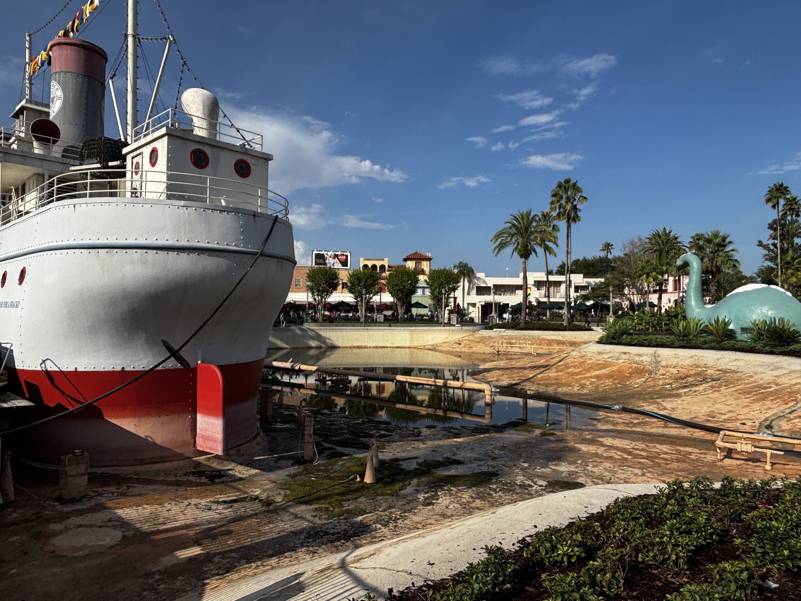 A white and red steamship-styled building resting on an empty lakebed, with algae staining the concrete and a hose and pipe running past its bow