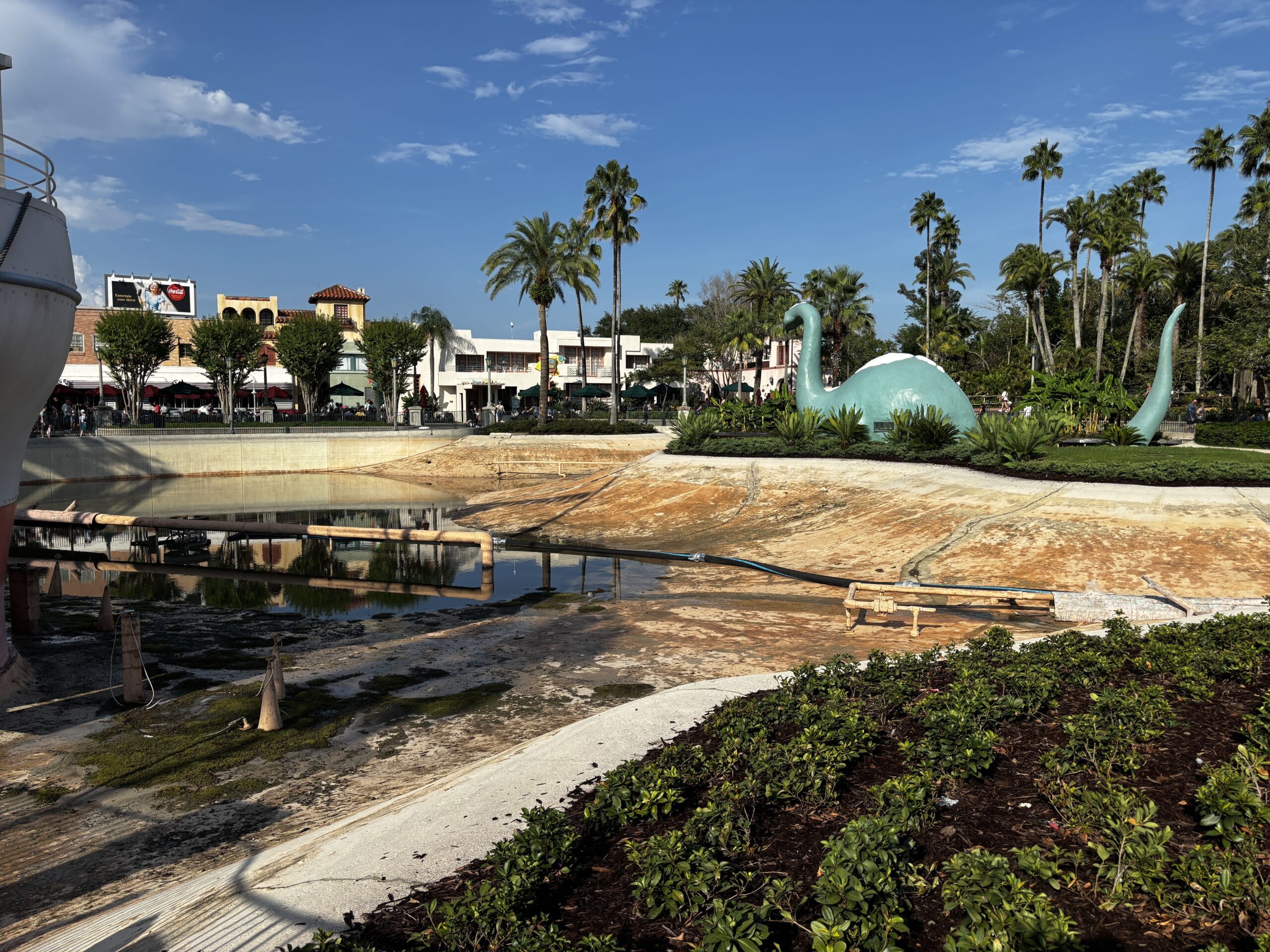 A wide view across the emptied Echo Lake basin toward Gertie the dinosaur building, with pumping equipment and a hose across the drained concrete and the remaining puddle