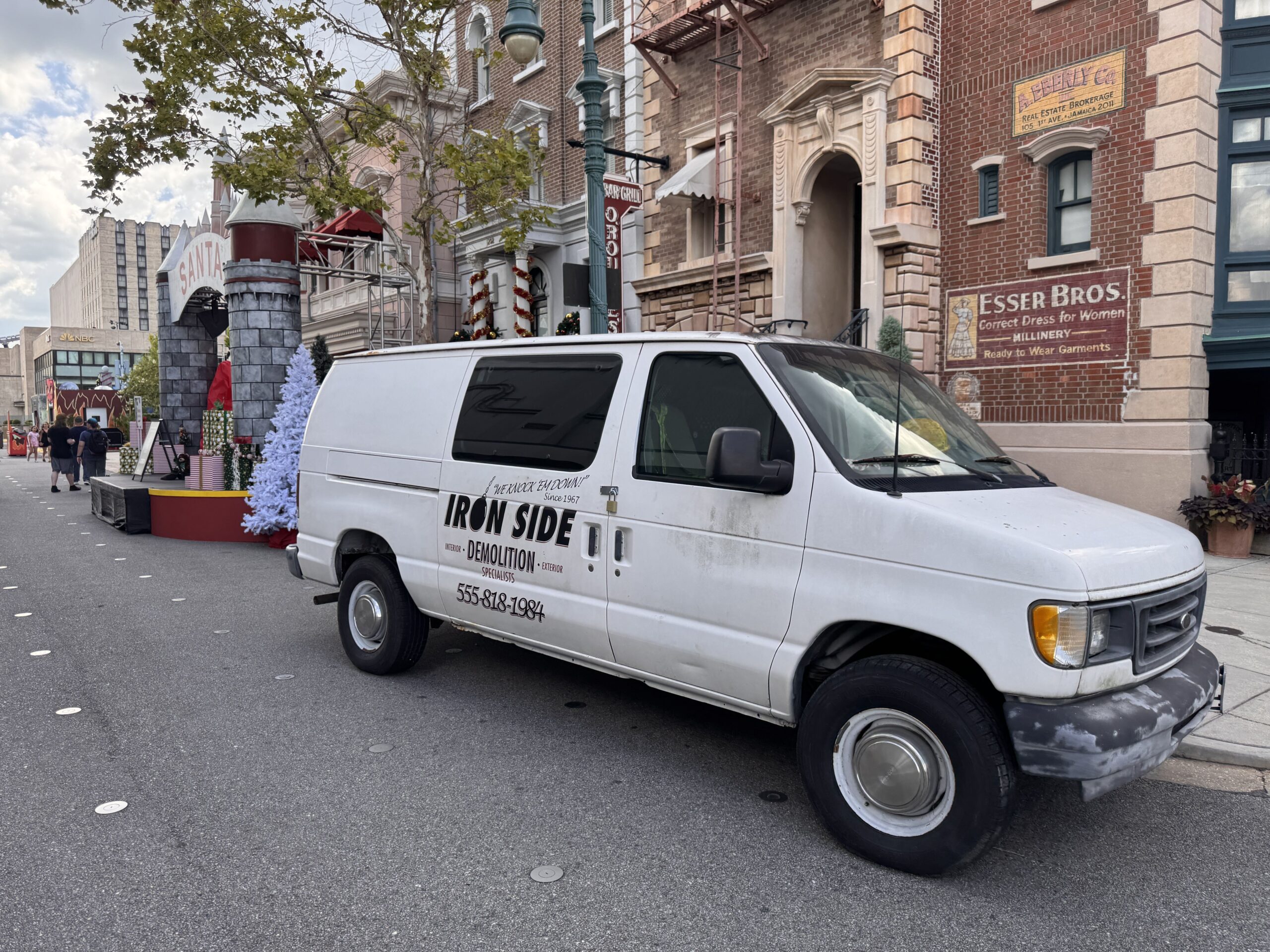 A weathered white cargo van lettered Iron Side Demolition Specialists parked on a theme park street, with the Santa arch and a white Christmas tree behind it