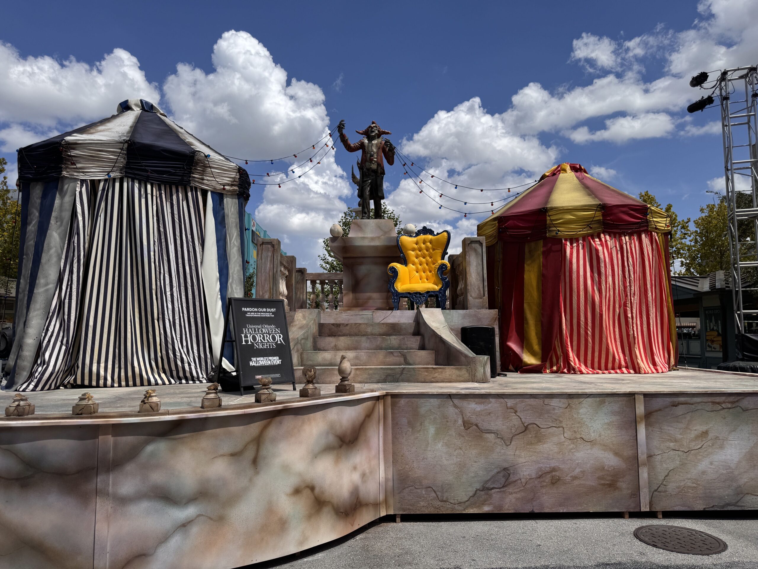 A weathered painted clown figure in a long ragged rust red coat under a matching pointed jester hat stands arms raised on a faux marble pedestal between a blue and white striped tent and a red and gold striped tent