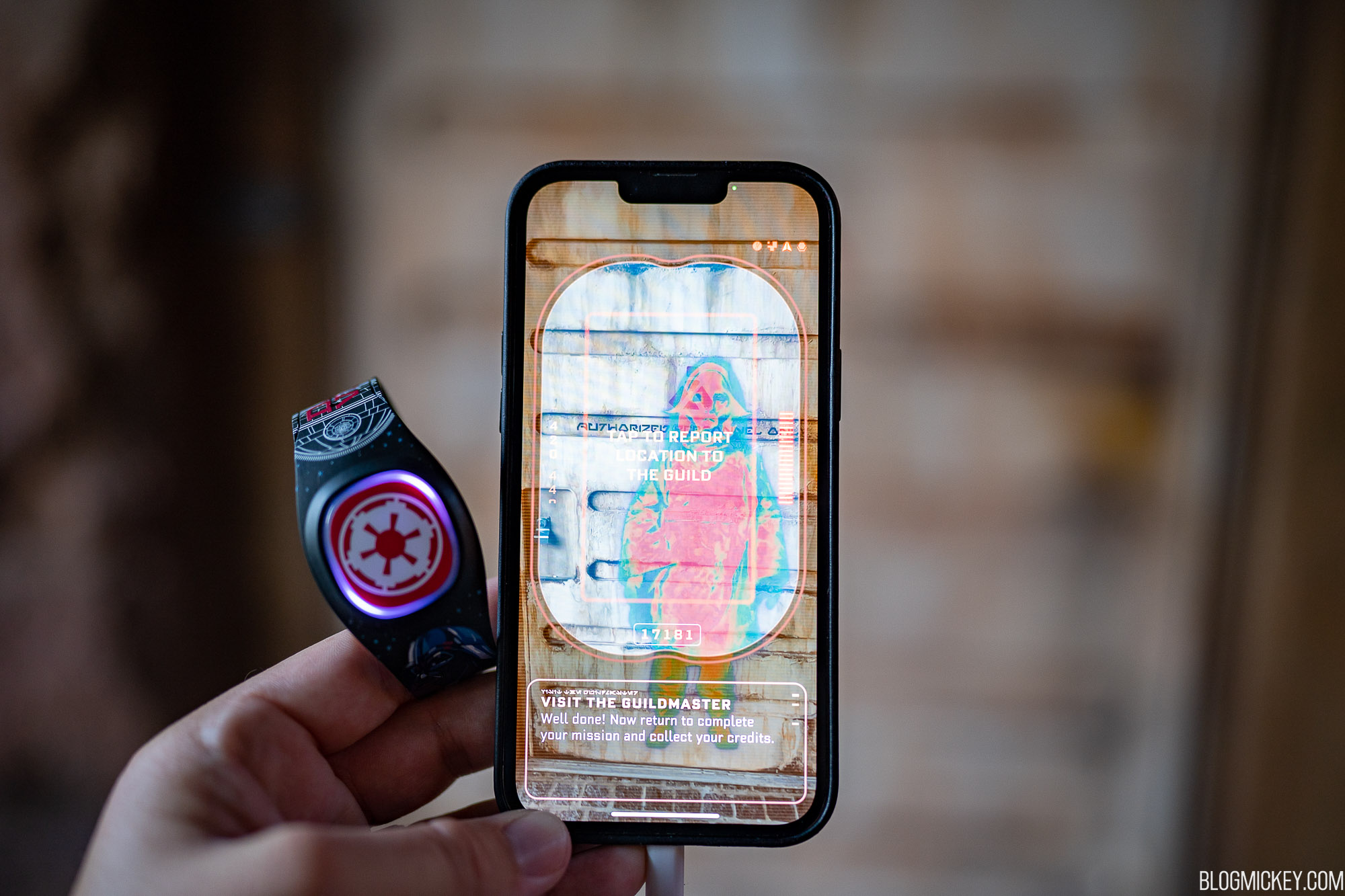 A hand holding a black Imperial crest MagicBand Plus with a glowing purple and red ring, next to a phone running the Batuu Bounty Hunters game