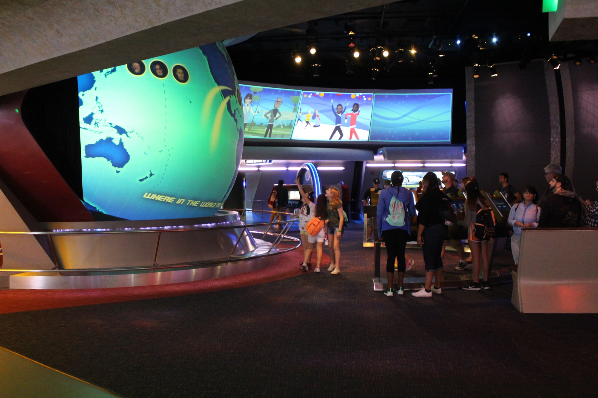 Guests standing in the Project Tomorrow post-show area beneath Spaceship Earth, gathered around a large illuminated globe screen showing world maps and the words WHERE IN THE WORLD, with video panels mounted overhead.