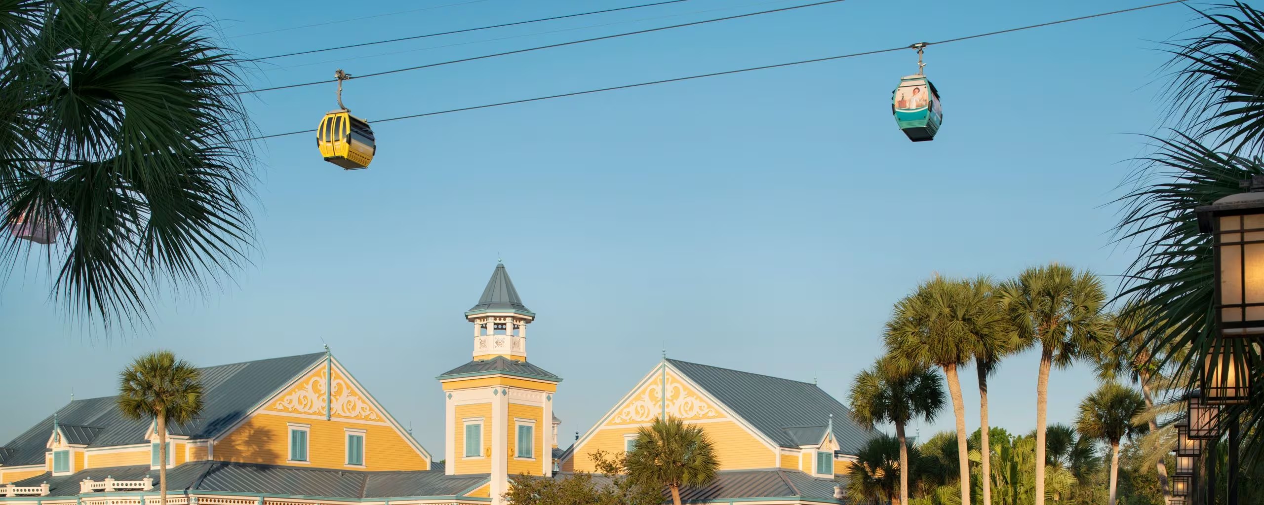 Yellow gabled buildings with white scrollwork and a cupola tower at Disney's Caribbean Beach Resort, with Disney Skyliner gondolas crossing overhead on a cable