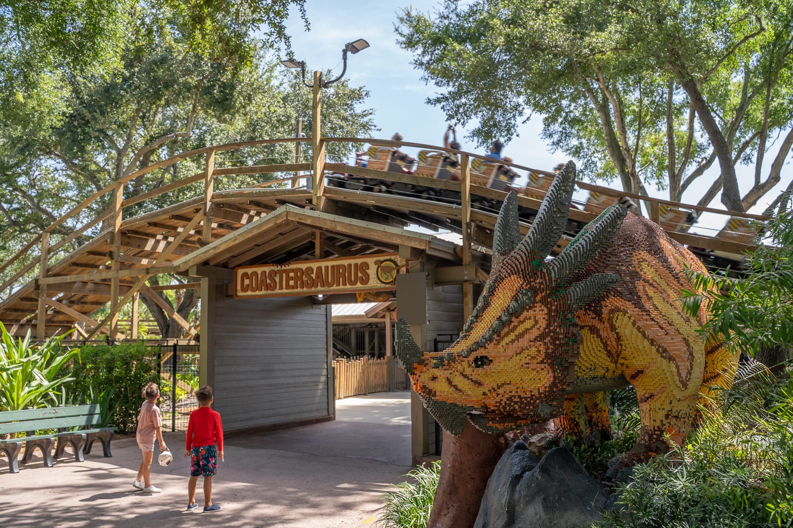 The Coastersaurus entrance building with a full train of riders on the wooden track above it and a large LEGO brick triceratops at right