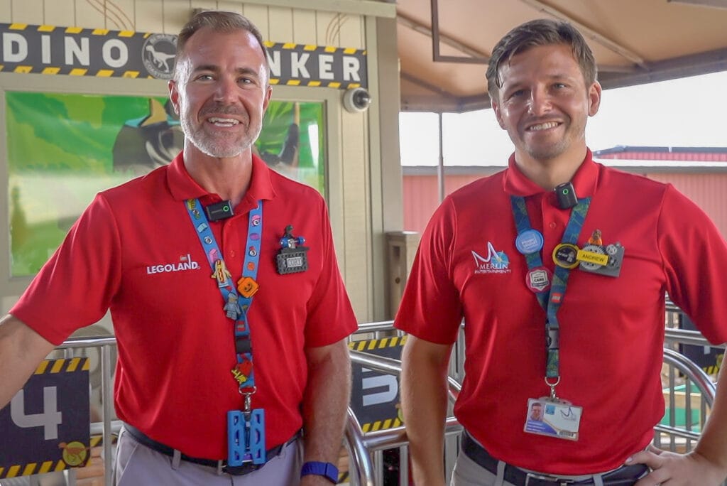 Two men in red polo shirts standing side by side on the ride platform in front of yellow and black DINO BUNKER signage and numbered row markers