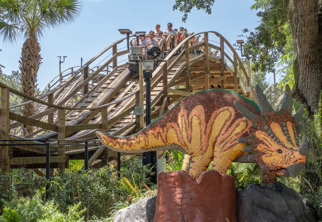 A full LEGO brick triceratops sculpture on a rock outcrop in the foreground with a train of riders cresting the wooden coaster track behind it