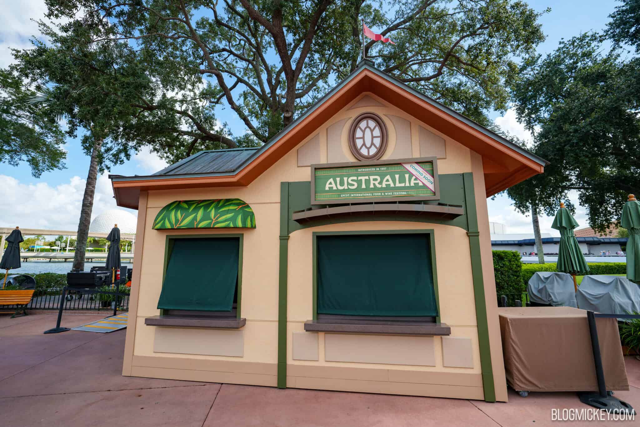 The Australia Global Marketplace kiosk at EPCOT, a tan cottage with an orange trimmed roofline and green posts, its green sign reading INTRODUCED IN 1997 above AUSTRALIA with a cream Coming Soon diagonal edged in maroon, both service windows shuttered in dark green canvas