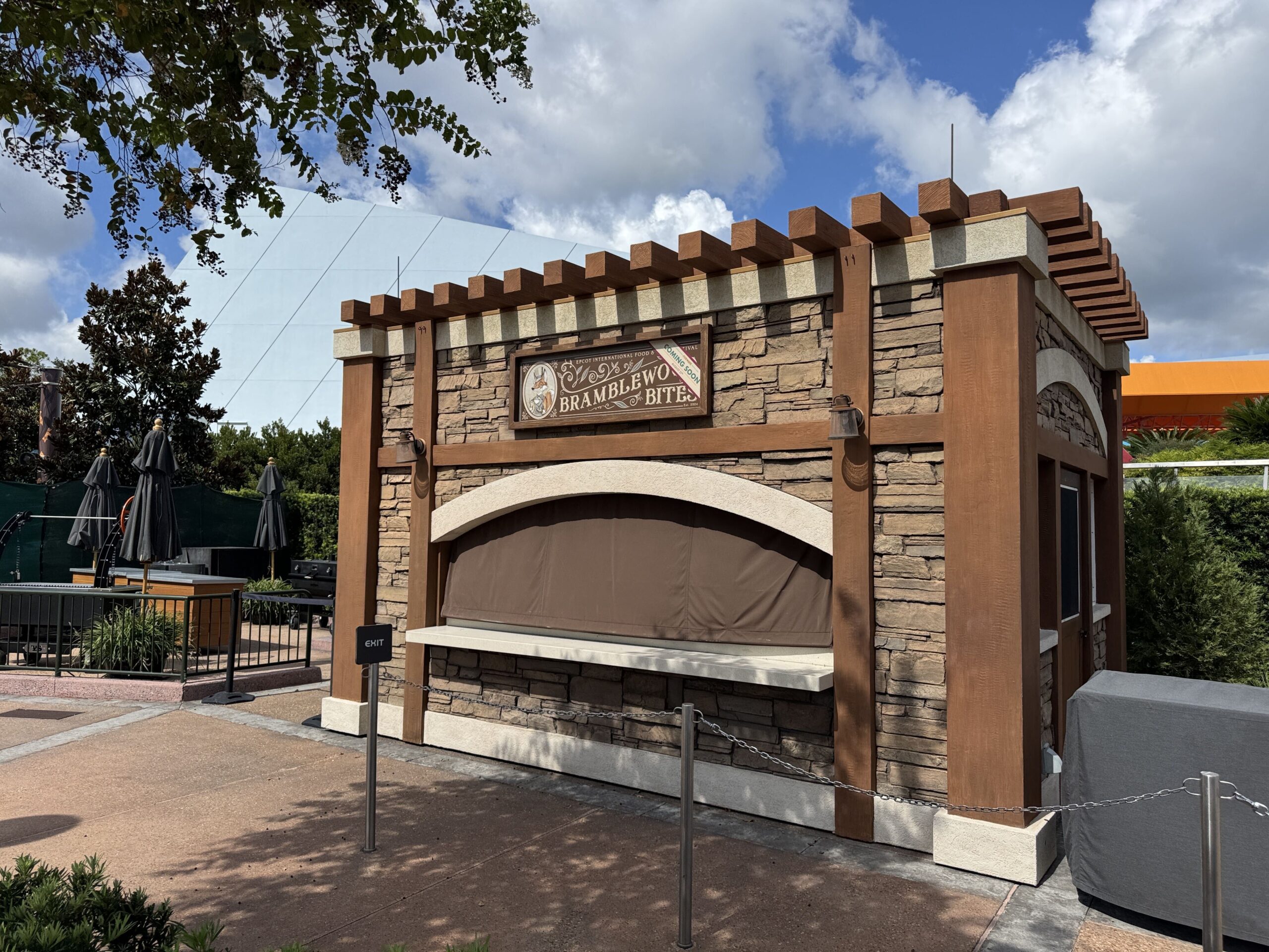 The Bramblewood Bites booth at EPCOT, a stone and timber structure with a wooden pergola cap, its framed sign showing an oval cameo of a top-hatted fox in a pale waistcoat beside the name, under a cream Coming Soon diagonal, the service window covered by a brown tarp and roped off with a chain