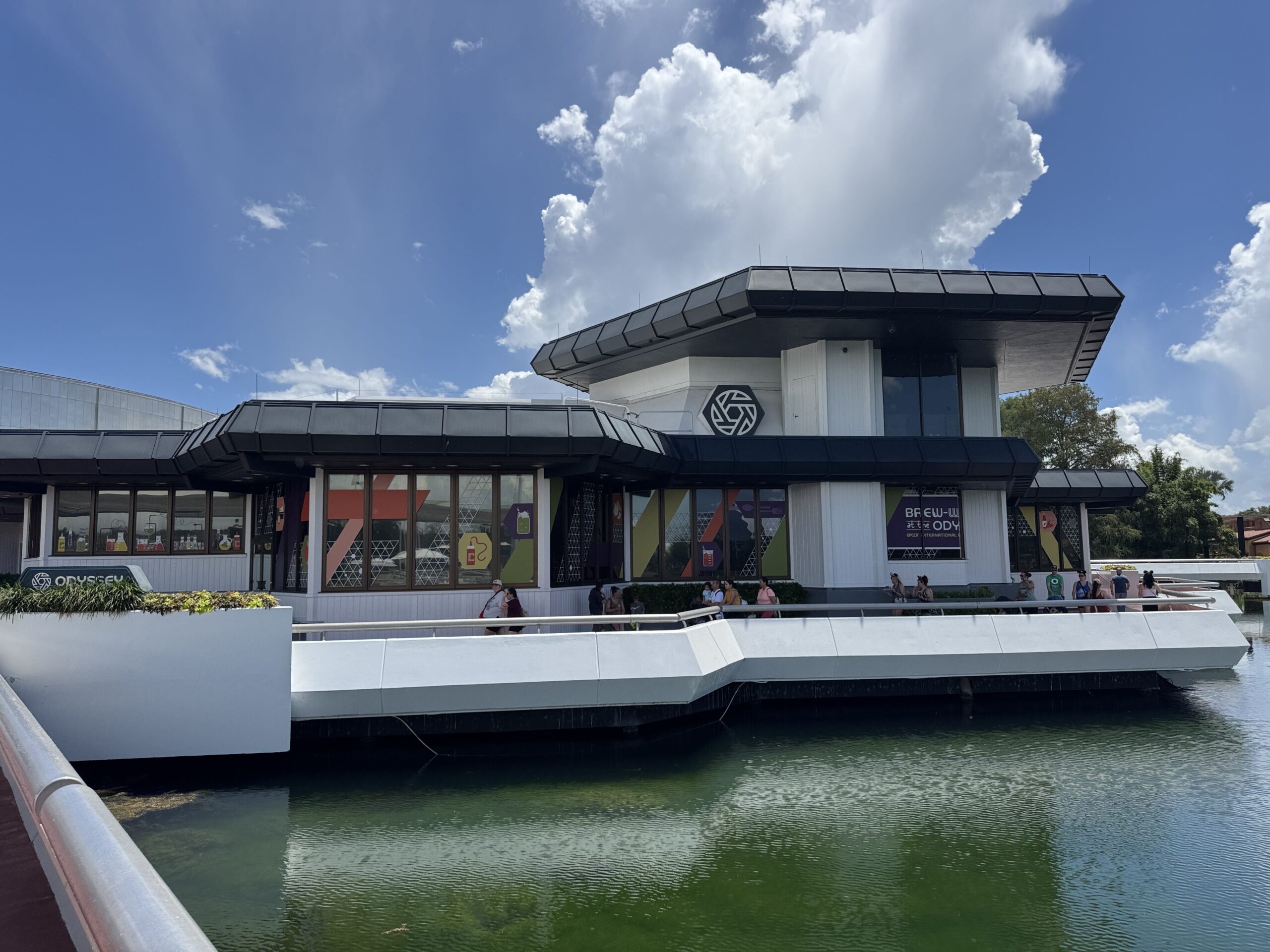 The Odyssey pavilion at EPCOT seen across the water, its windows covered in lab themed decals of beakers and flasks in orange, purple and green, a purple Brew-Wing at the Odyssey banner on the near corner and the green ODYSSEY sign at left, with guests walking the white bridge in front of it