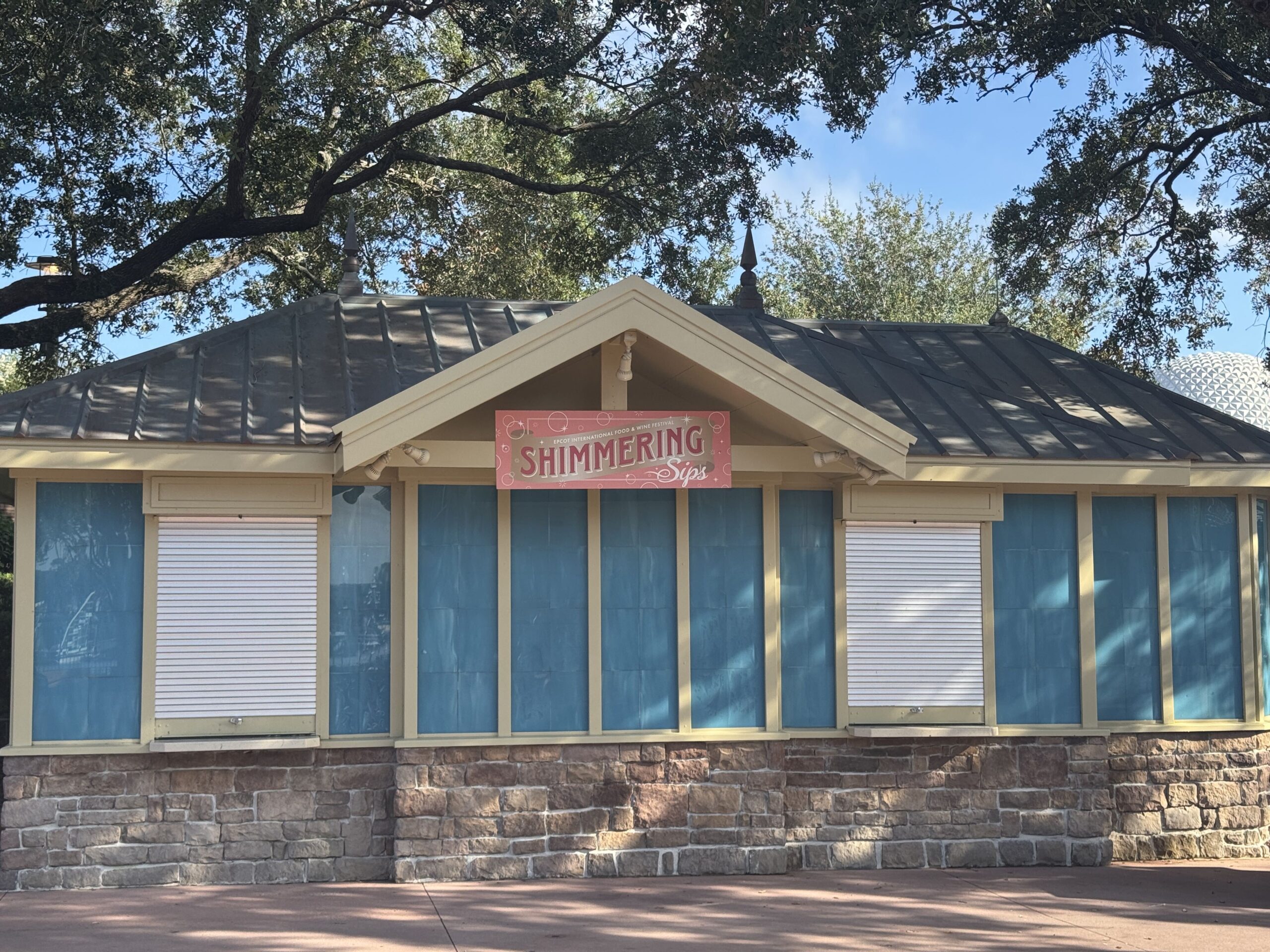 The Shimmering Sips kiosk at EPCOT, a cream and teal building with a dark metal hipped roof and a stone base, its coral pink sign reading SHIMMERING Sips hung under the gable, its service windows rolled shut