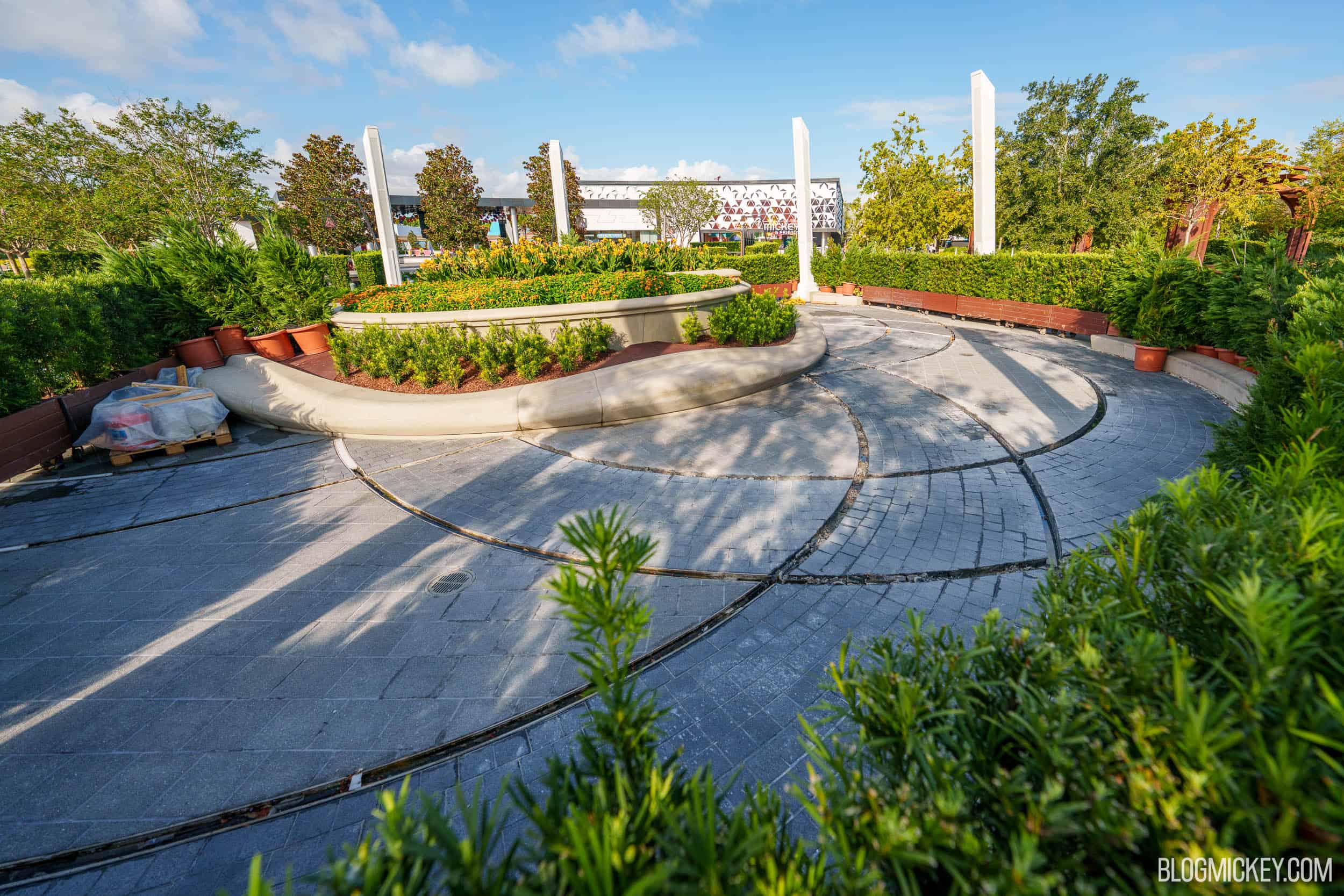 The same EPCOT plaza with dark empty channels cut into the paver walkways where the lighting strips have been removed, a pallet of wrapped material at left