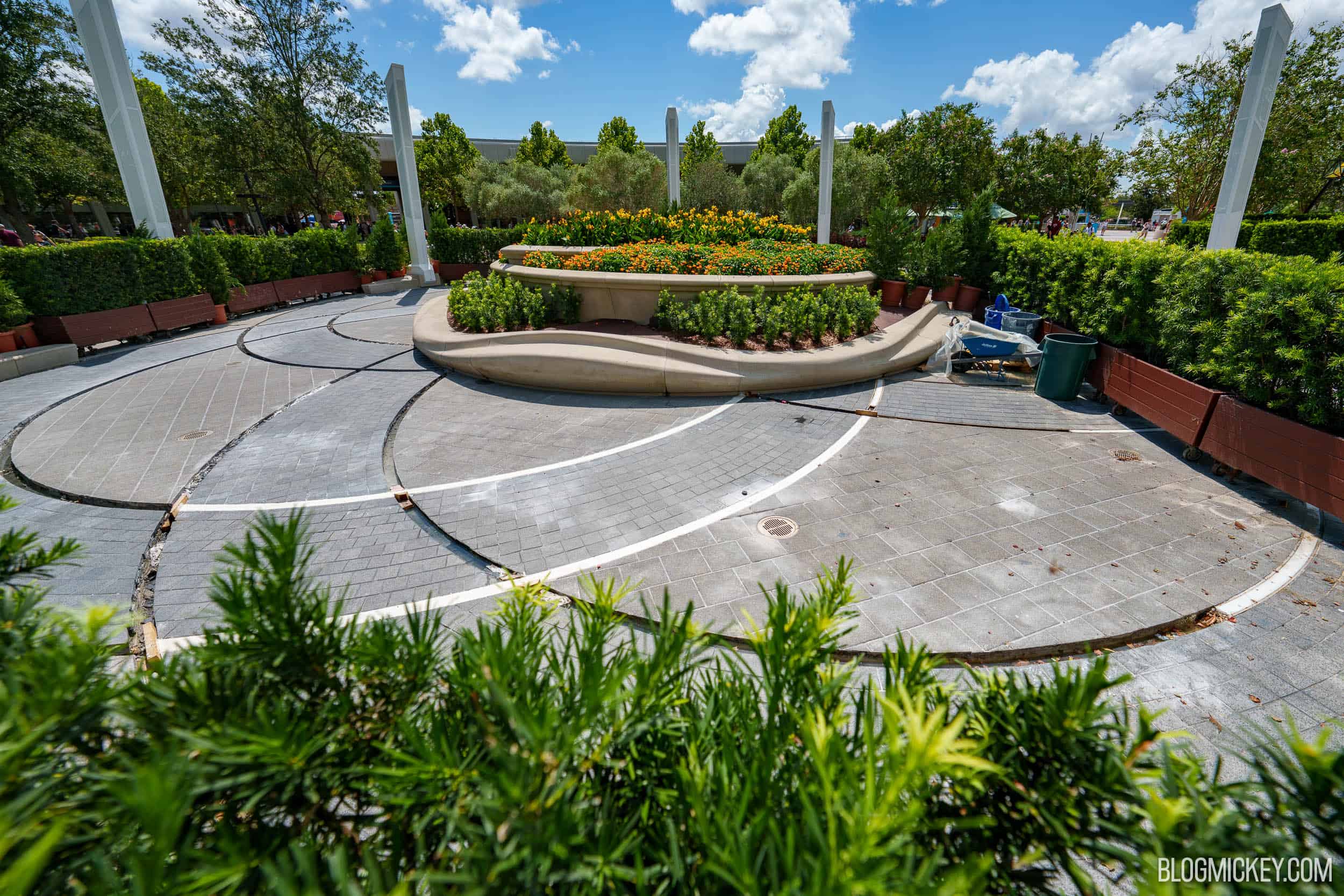 Curved walkways in EPCOT's World Celebration Gardens with pale concrete fill tracing the curves where lighting strips used to sit, a wheelbarrow and buckets staged beside the central planter