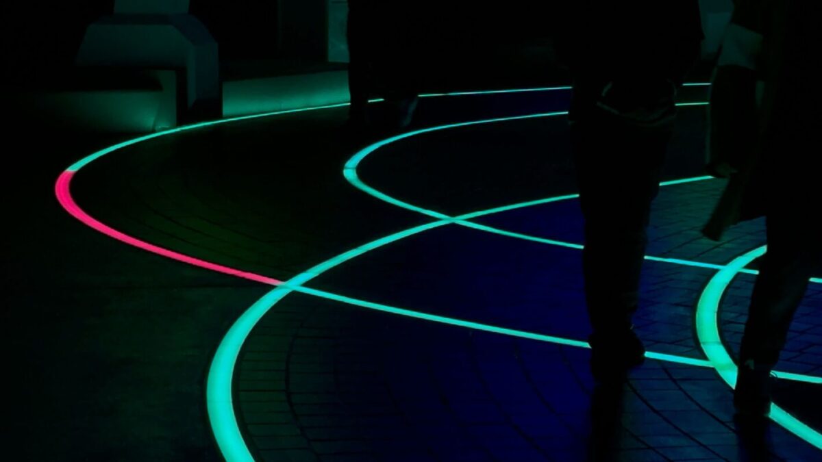 The in-ground lighting strips glowing in curved lines across dark pavement at night, most of them teal while one curve on the left glows pink, with guests walking past