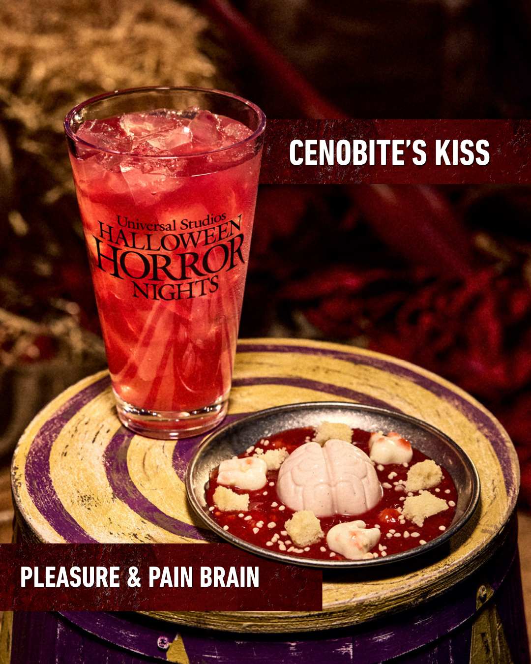 A red iced drink labeled Cenobite's Kiss in a Halloween Horror Nights glass next to a dark plate labeled Pleasure and Pain Brain holding a pale molded brain in red sauce with white pieces around it, set on a purple and gold striped barrel top