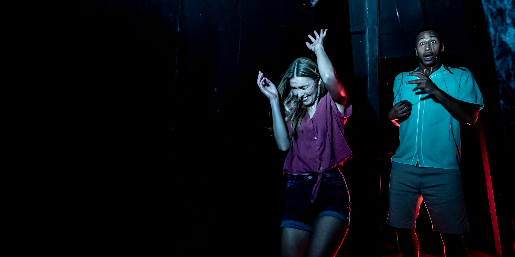 Two guests reacting in fear in a dark haunt corridor lit in teal and red, a woman in a purple tied-front shirt and denim shorts with both hands thrown up beside a man in a light blue shirt and grey shorts, wide-eyed with his hands raised