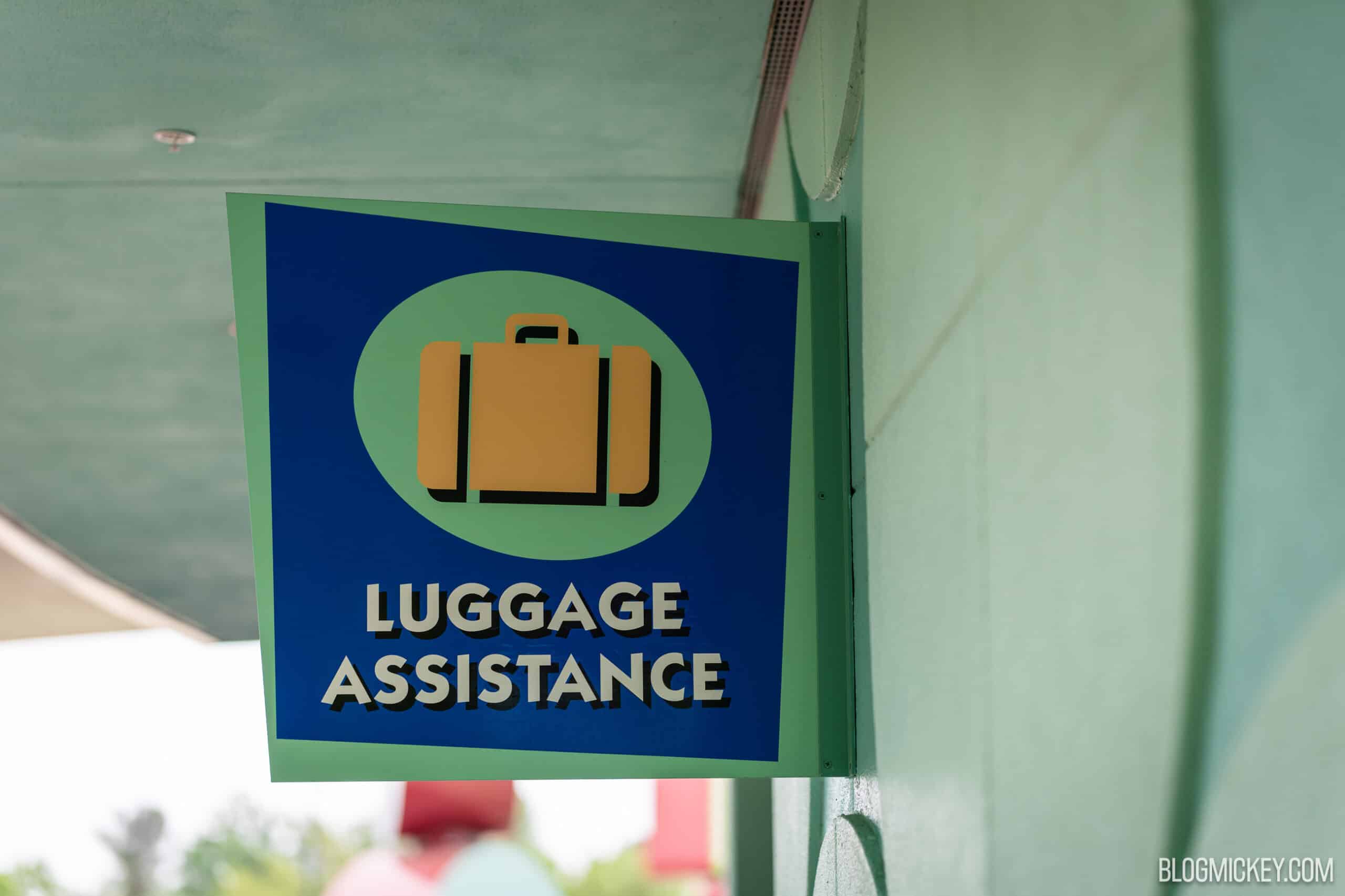 A blue Luggage Assistance sign with an orange suitcase icon on a green oval, mounted on a mint green resort column under a covered walkway