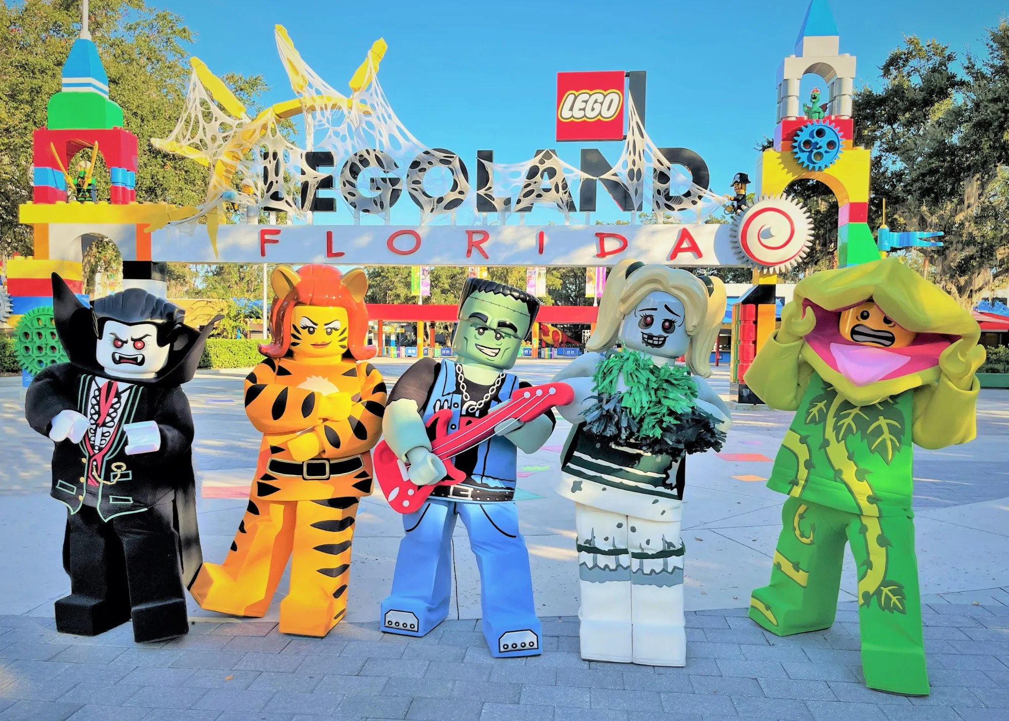 Five walkaround LEGO monster characters stand in daylight in front of the LEGOLAND Florida entrance archway draped in fake spiderwebs, including a vampire in a black cape, an orange tiger character, a green Frankenstein rocker with a red guitar, a zombie cheerleader with green and black pom poms and a green plant monster