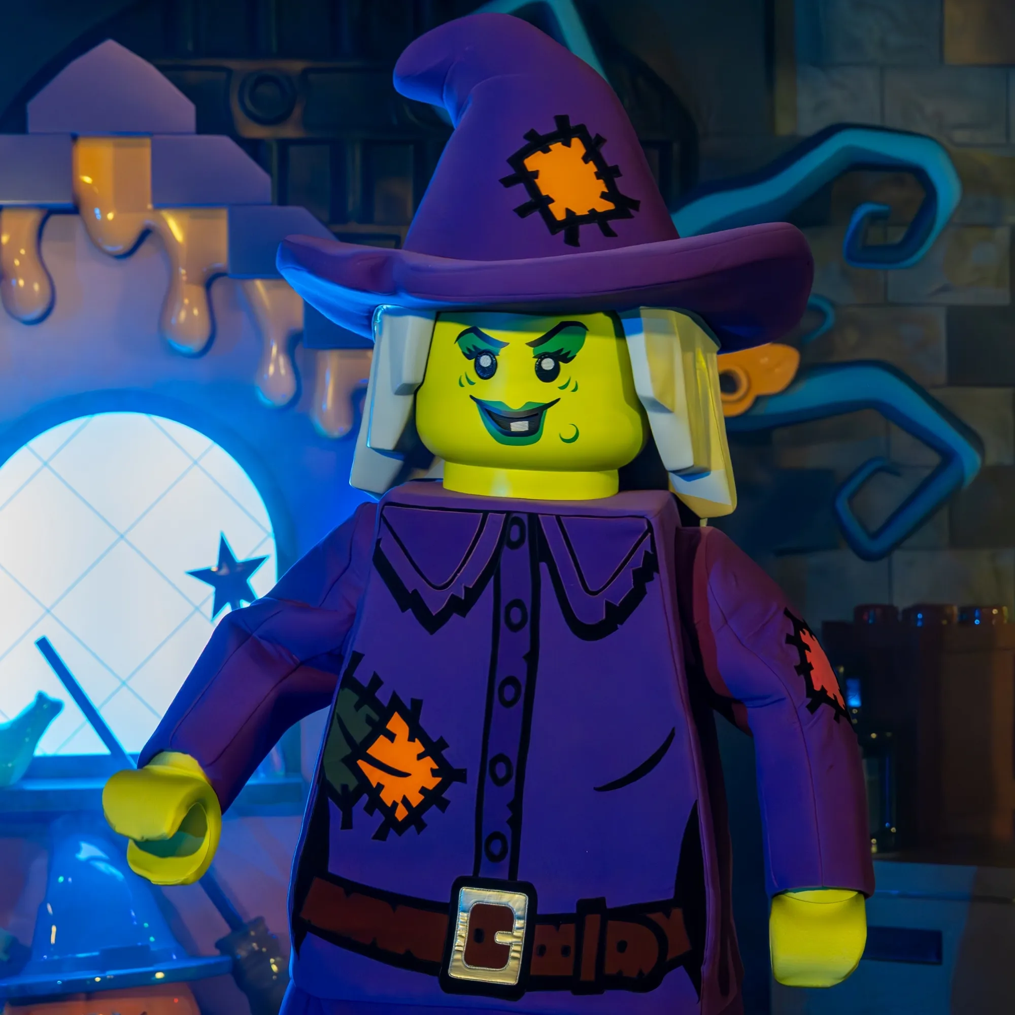 Close up of the LEGO Wacky Witch walkaround character, a lime green minifigure face under a patched purple witch hat, wearing a patched purple coat with a brown belt and gold buckle, lit in blue against a themed set with a glowing round lattice window behind her