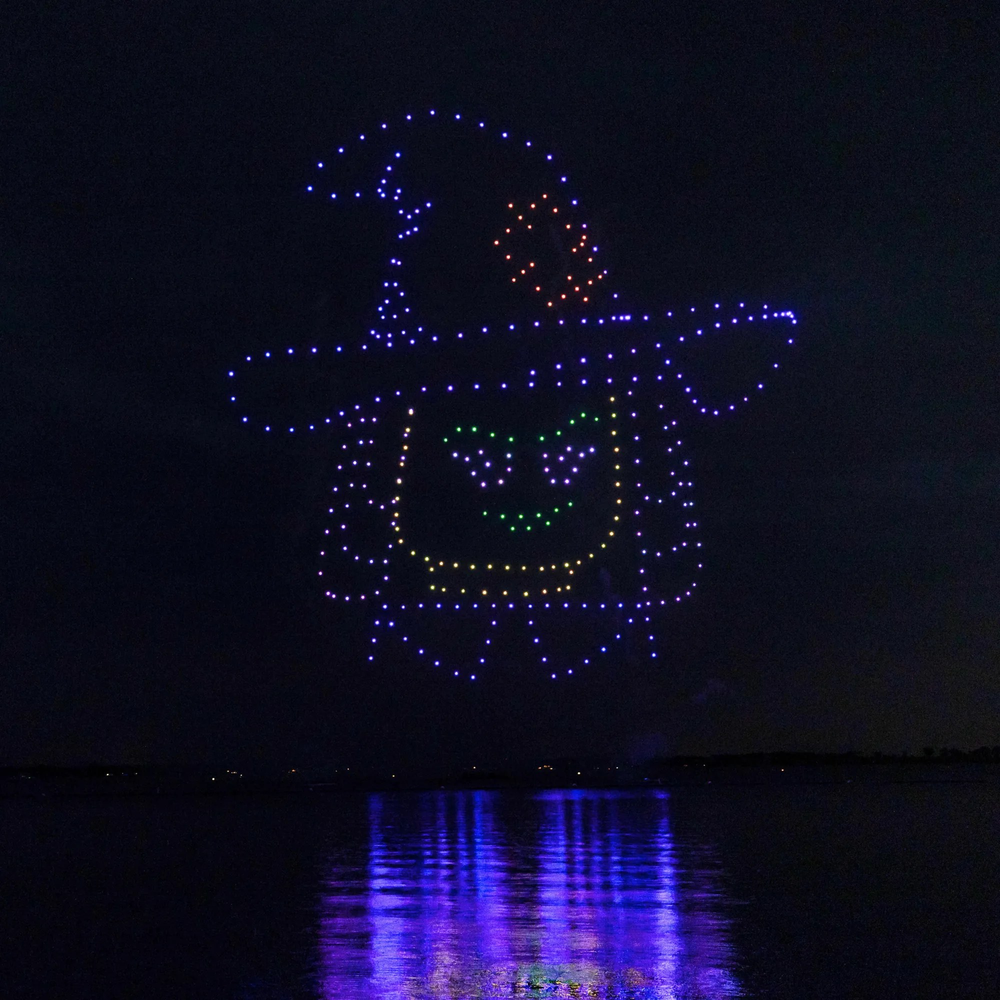 Hundreds of drones form a giant LEGO witch minifigure head, its eyebrows and grin picked out in green, under a wide brimmed pointed hat with an orange patch, against a black night sky, reflected in the water below