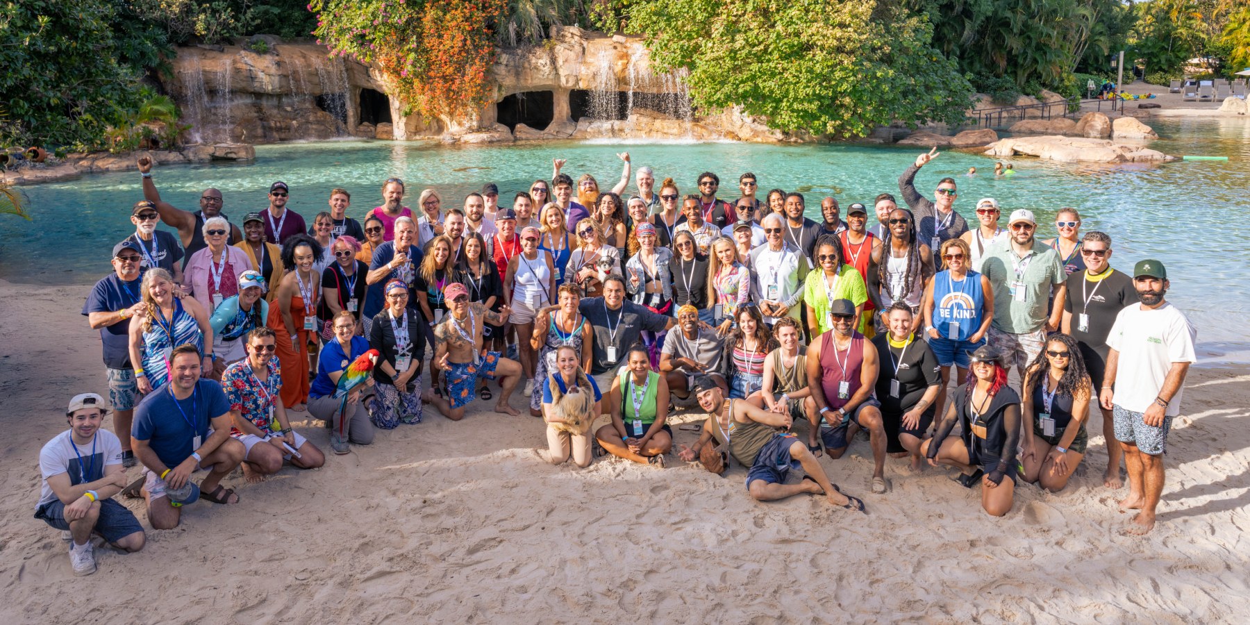 A large posed group of dozens of people wearing lanyards on a white sand beach in front of a turquoise lagoon and a rock waterfall grotto