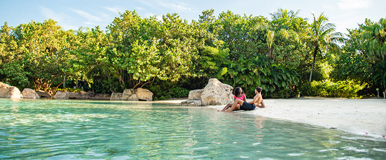 Two people sitting at the edge of a calm turquoise beach lagoon backed by dense green trees and palms