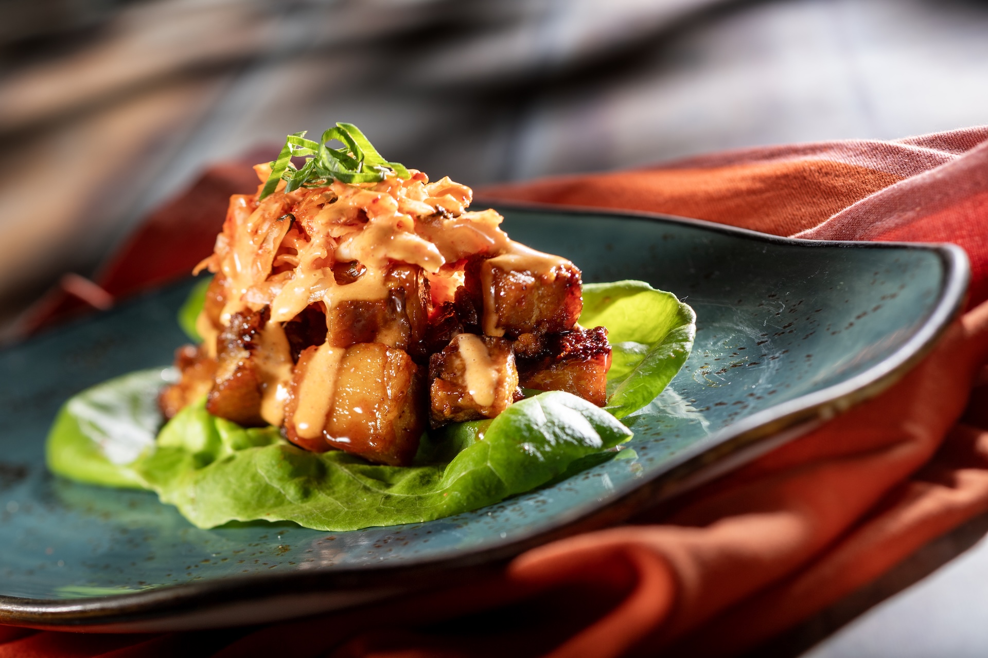Glazed cubes of pork belly stacked on a butter lettuce leaf, topped with kimchi slaw, a spicy orange aioli drizzle and sliced scallions, plated on a dark teal dish over a rust colored cloth