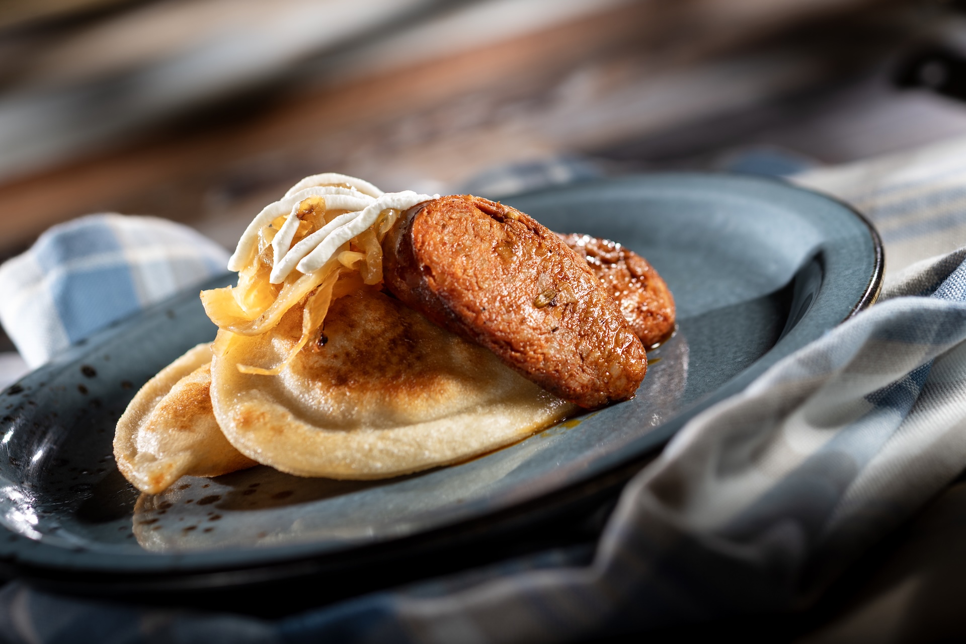 Two golden pan-fried potato pierogi on a dark blue plate, topped with caramelized onions and a drizzle of sour cream, with a browned length of kielbasa resting alongside and a blue checked cloth in the background