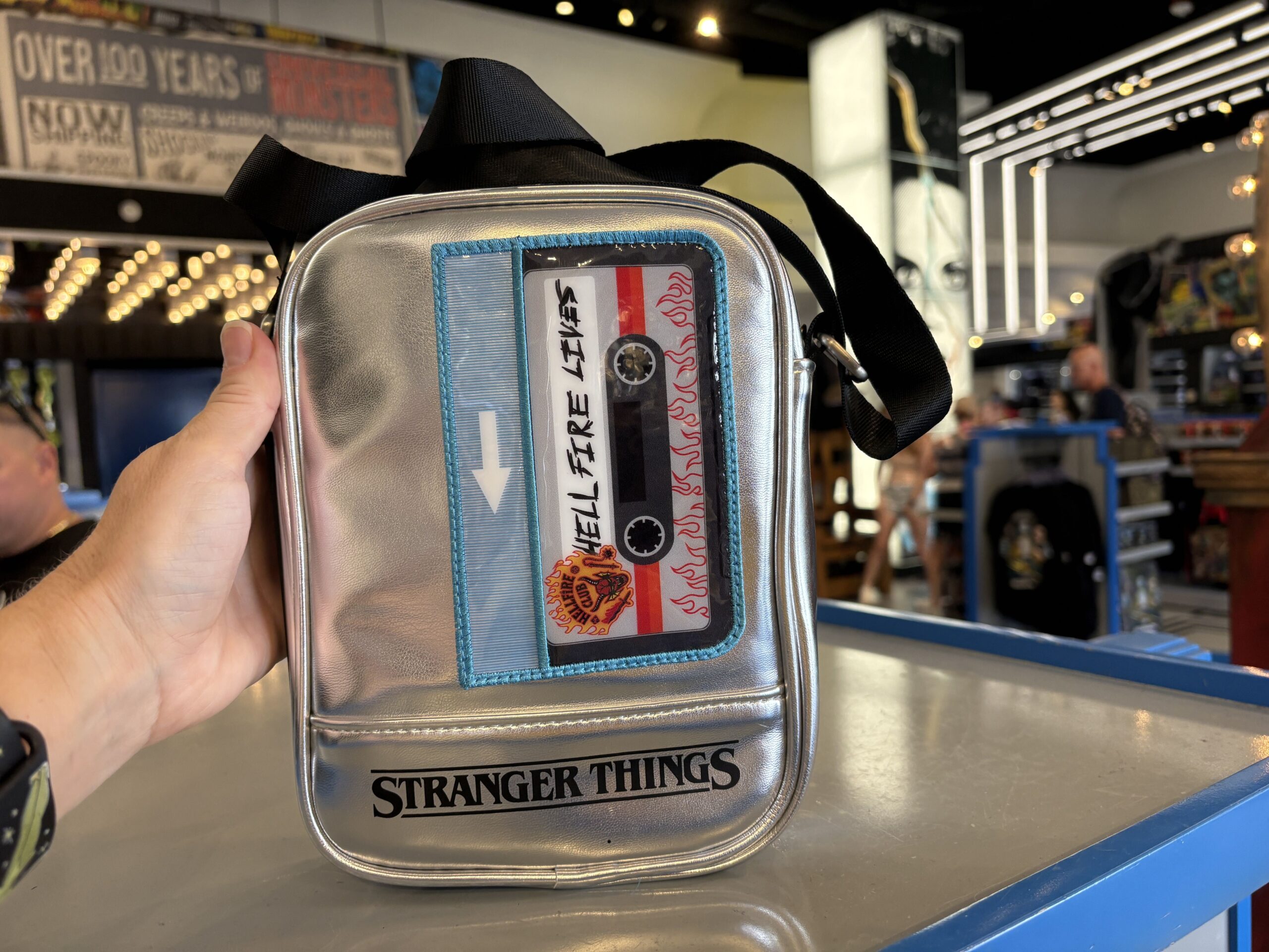 A hand holding a metallic silver crossbody bag shaped like a personal cassette player with a clear window over a Hell Fire Lives tape