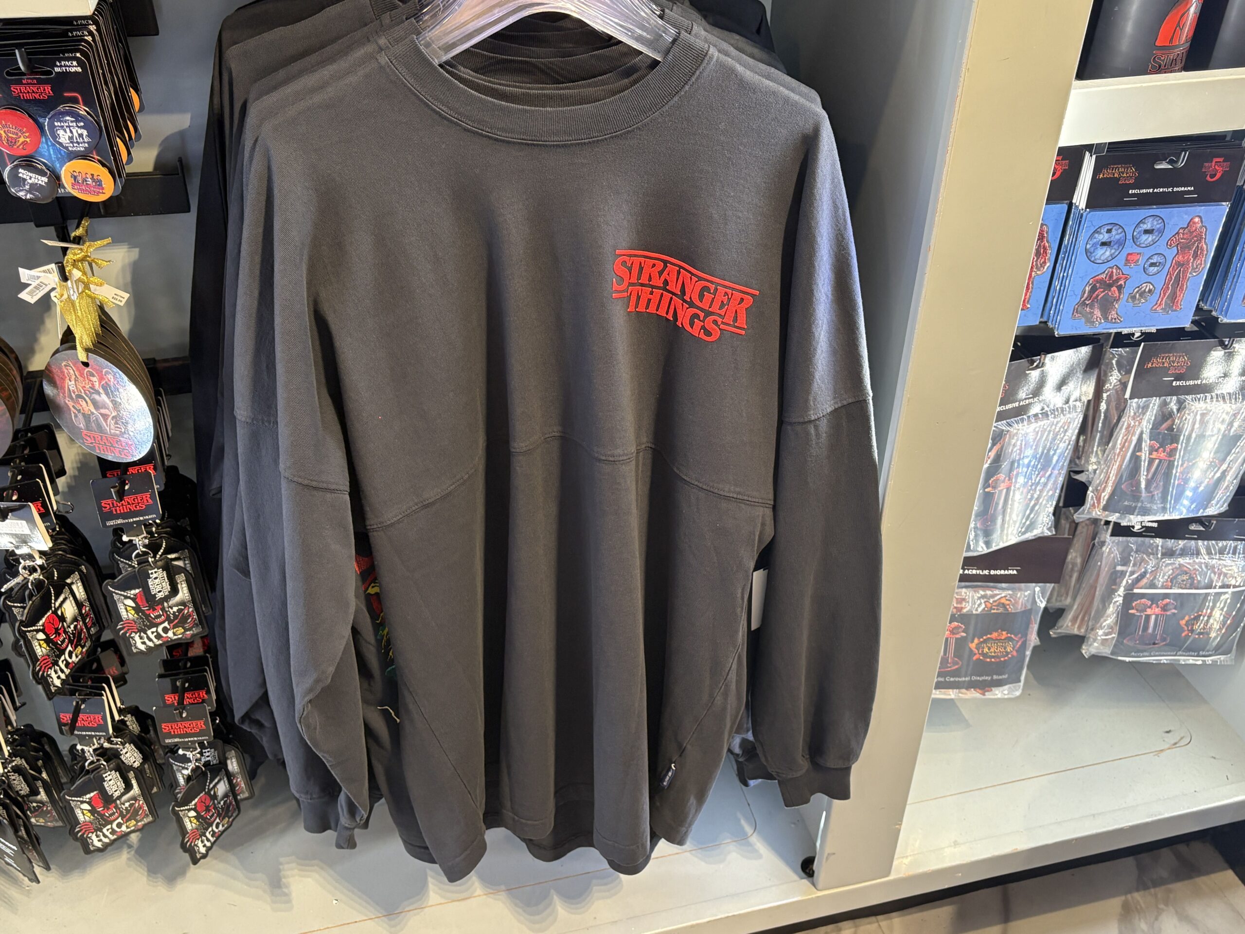 A long-sleeved spirit jersey on a hanger with a red raised Stranger Things logo on the left chest, button packs and acrylic dioramas on racks either side