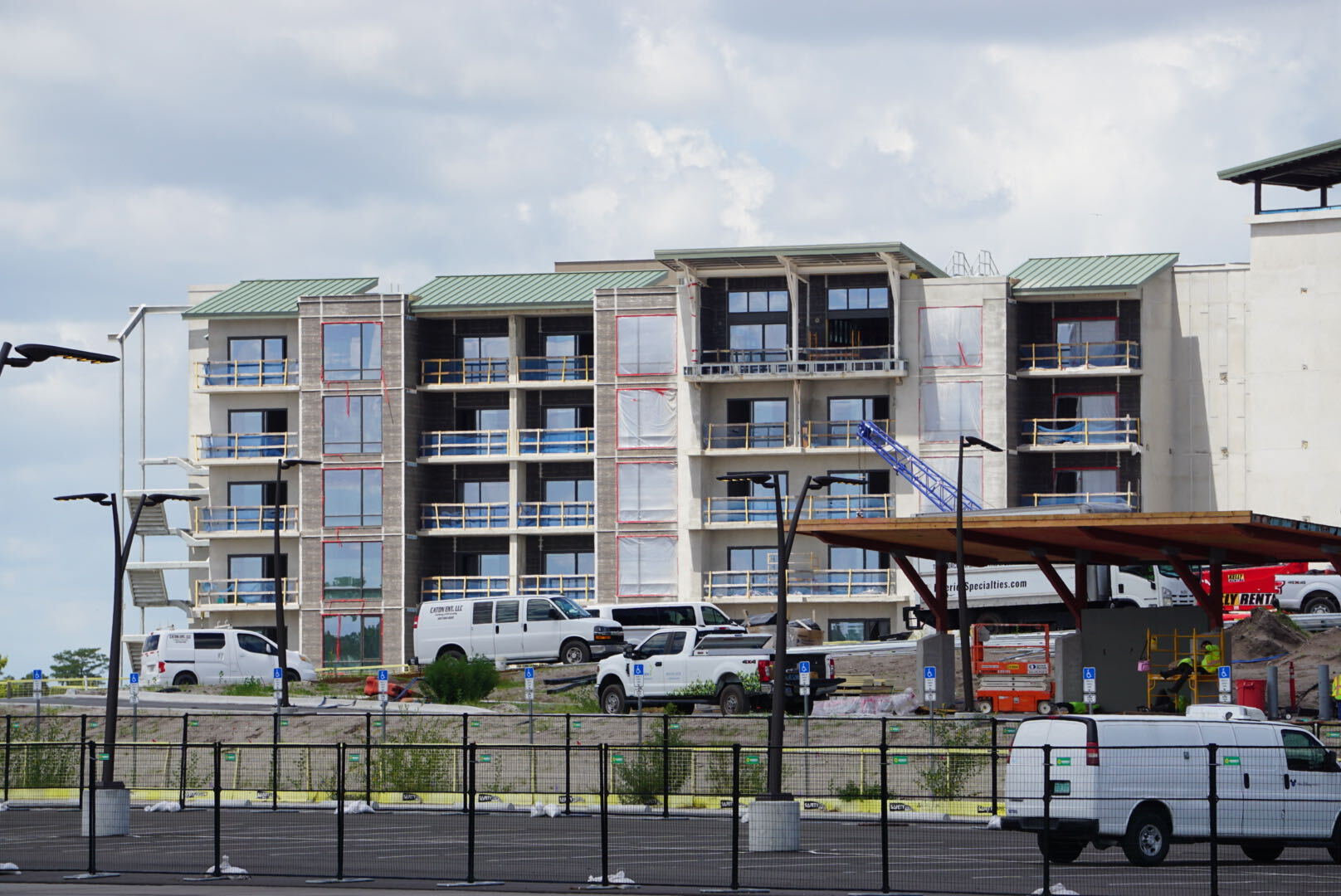 Ground level view of a five-story guest wing with green metal roofing, balconies behind yellow safety rails, and contractor vehicles parked in front
