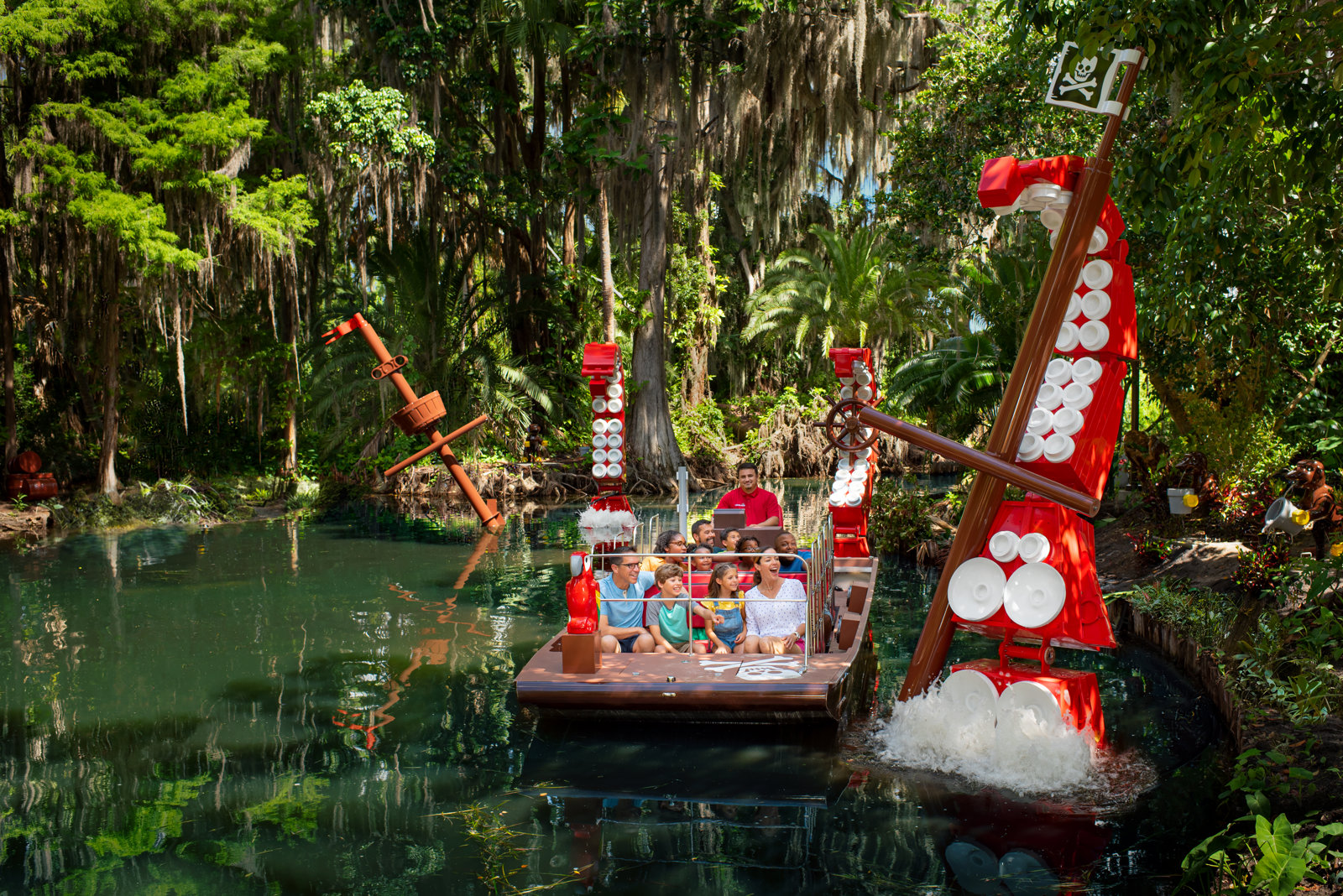 A flat-bottomed boat carrying a family and an operator drifting past a large red and brown LEGO pirate ship rising out of a green canal