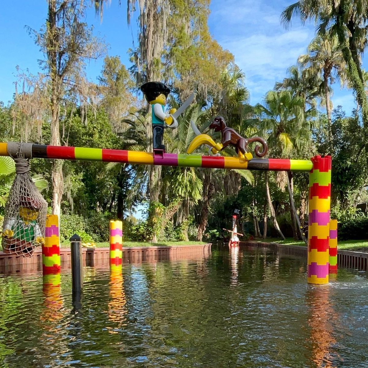 A LEGO pirate minifigure holding a cutlass facing a brown LEGO monkey with a banana, both standing on a colorful striped beam spanning a canal