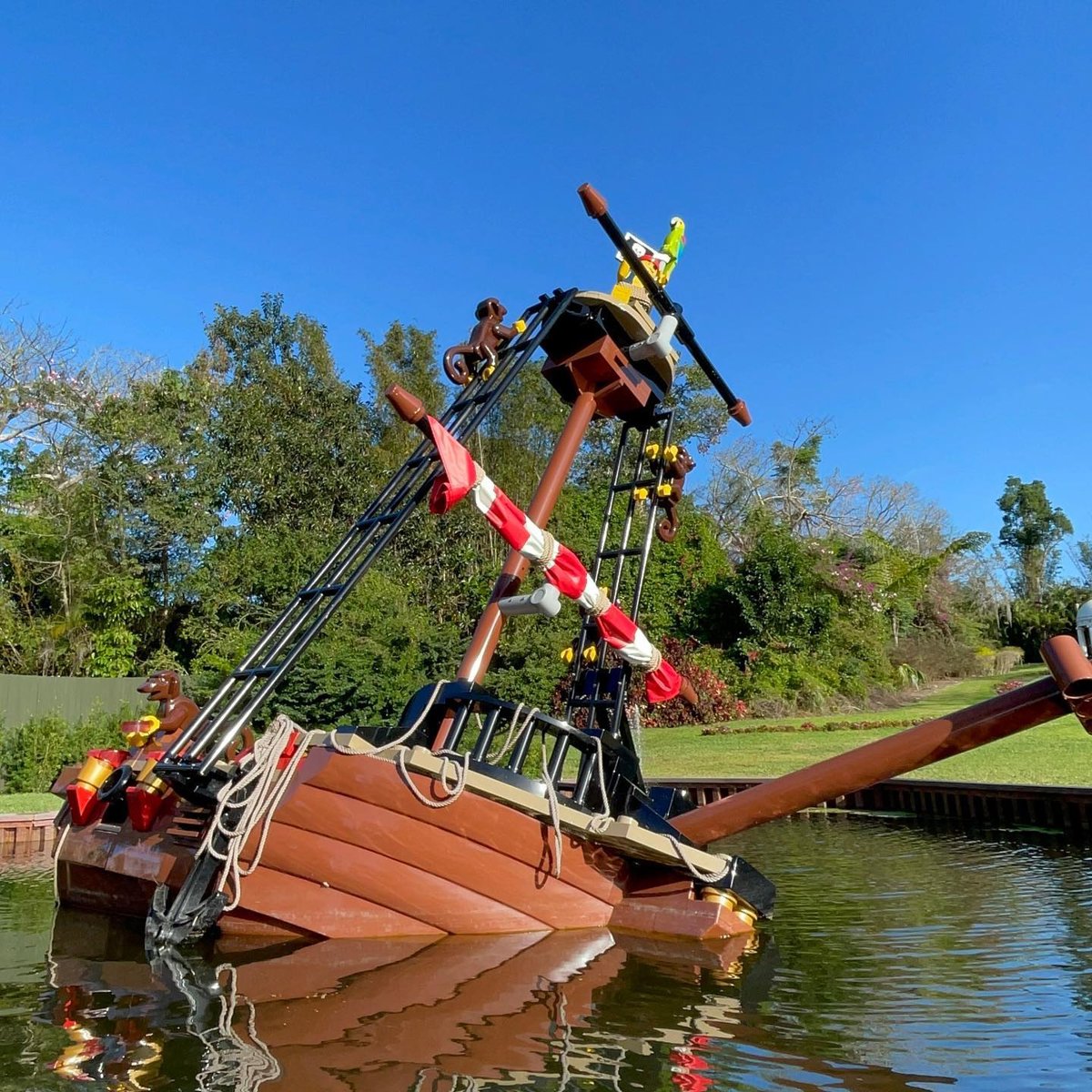 A half sunk LEGO pirate shipwreck in a canal with black rigging ladders, furled red and white sails, a crow nest with a LEGO parrot perched beside it and monkeys climbing the rigging ladders