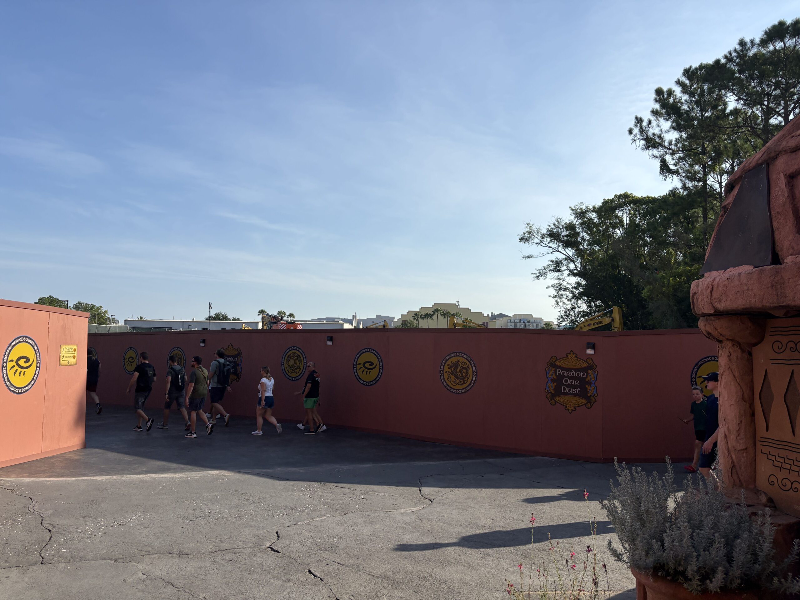 Guests walk past a long rust red construction wall decorated with gold sun and serpent medallions and two Pardon Our Dust placards, with several yellow excavator booms rising above the wall, one of them marked Kobelco