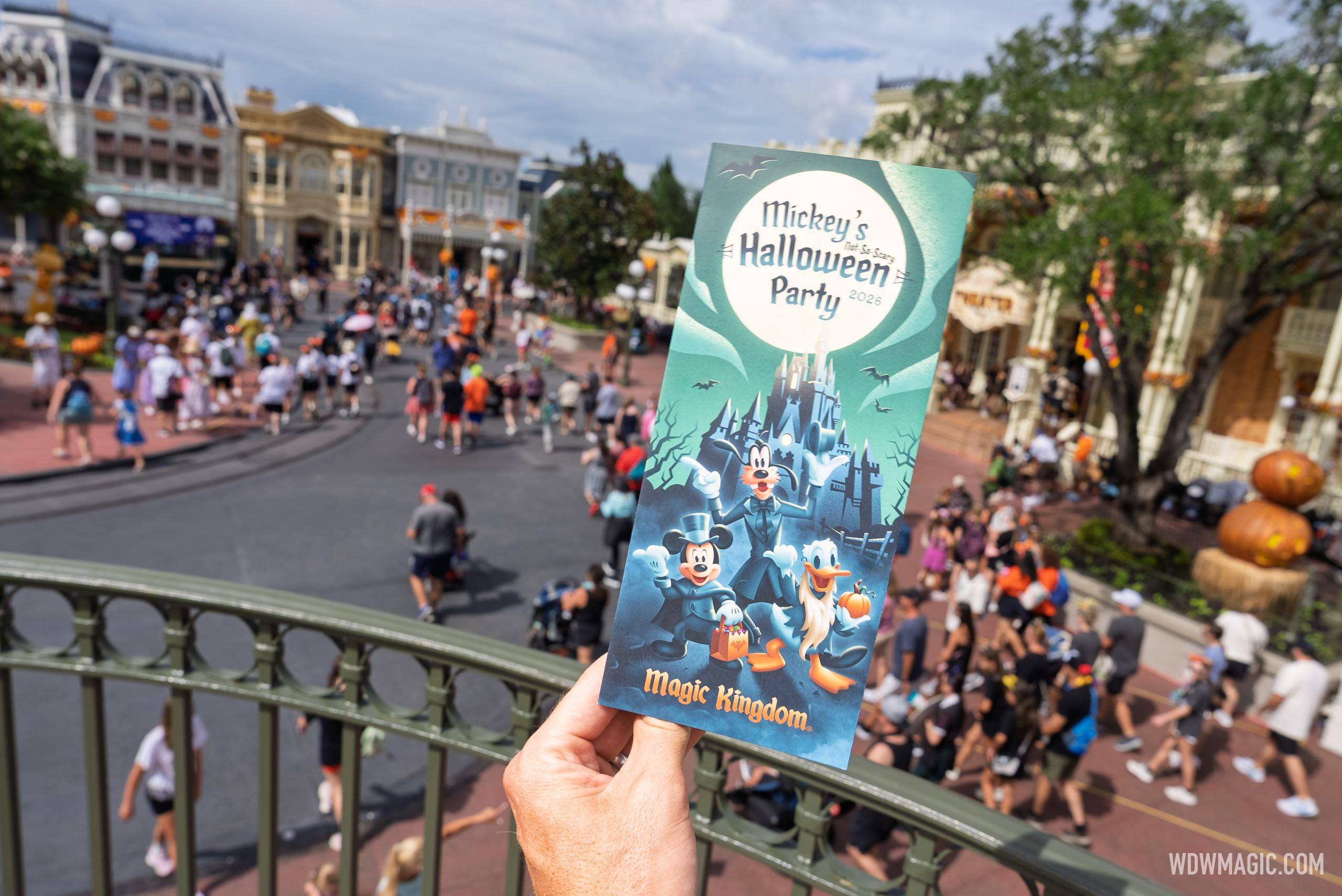 A hand holds up the teal 2026 party guide map showing Mickey in a top hat, Goofy in tails and Donald holding a pumpkin under a full moon, above a crowded Main Street