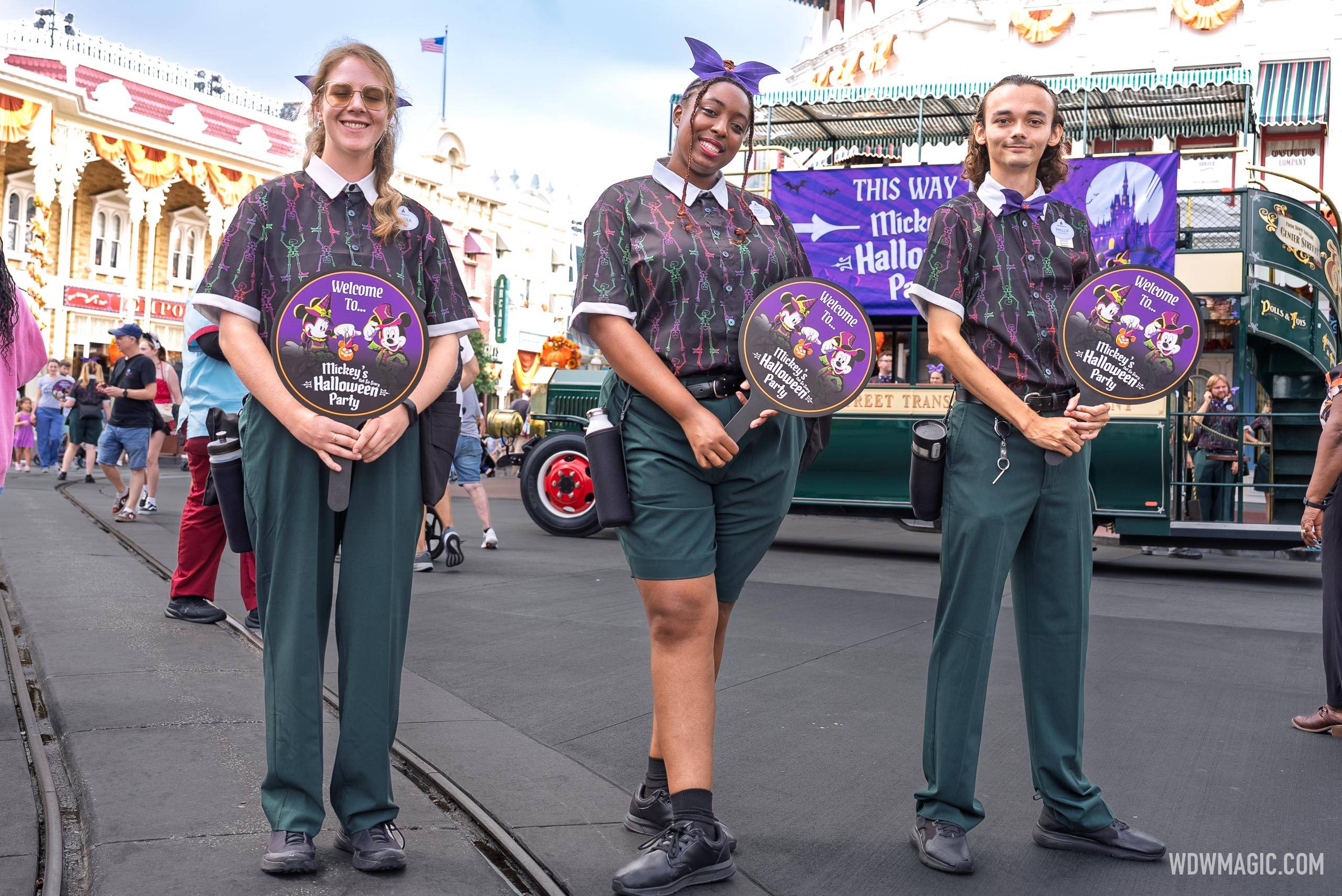 Three cast members in dark shirts printed with neon skeletons, two in green trousers and one in green shorts, hold round Welcome To Mickey's Not-So-Scary Halloween Party paddle signs on Main Street, with the green trolley and a purple party banner behind them