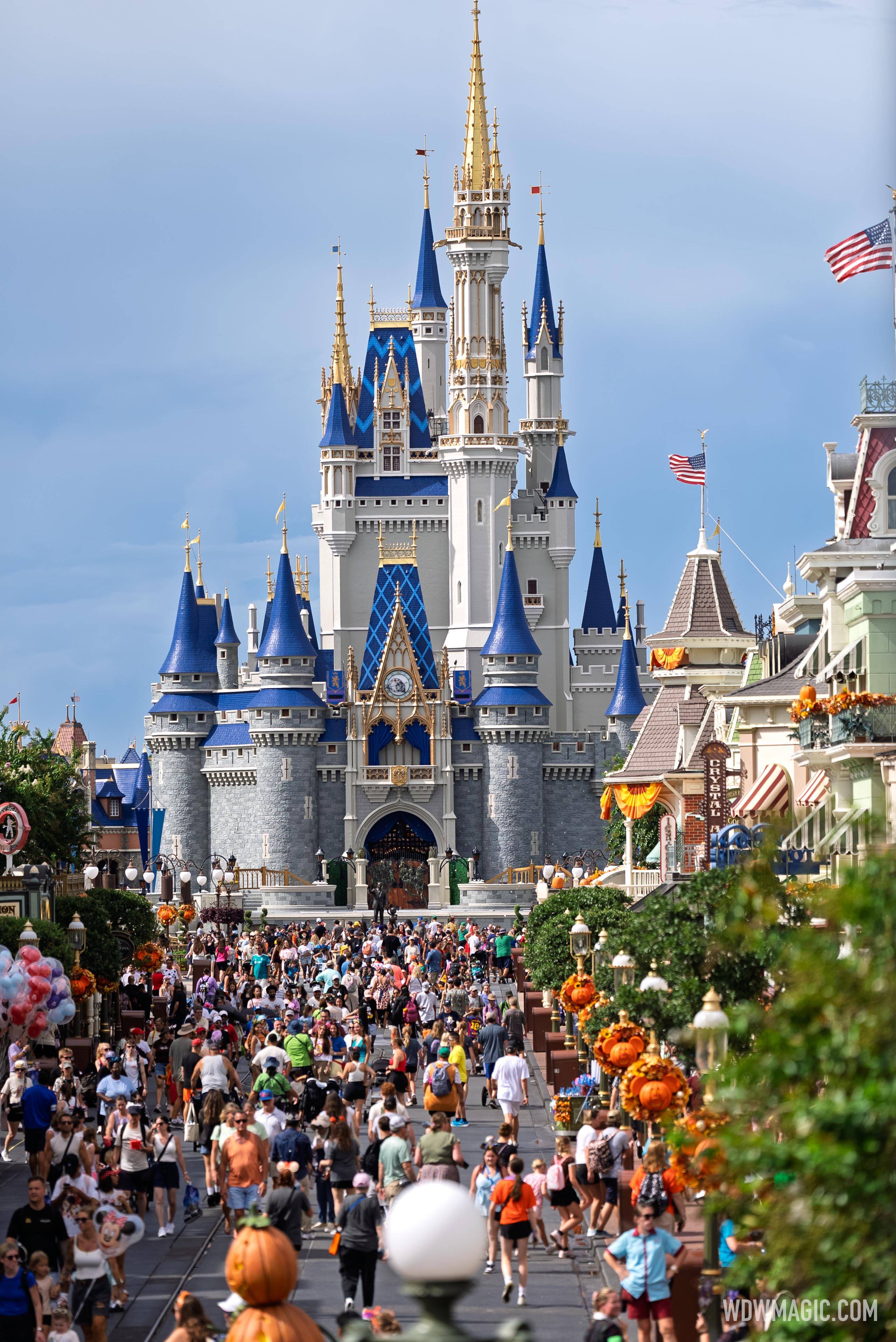 A long lens view down a crowded Main Street to Cinderella Castle, with orange pumpkin and fall foliage wreaths on the lamp posts and orange drapery on the shops