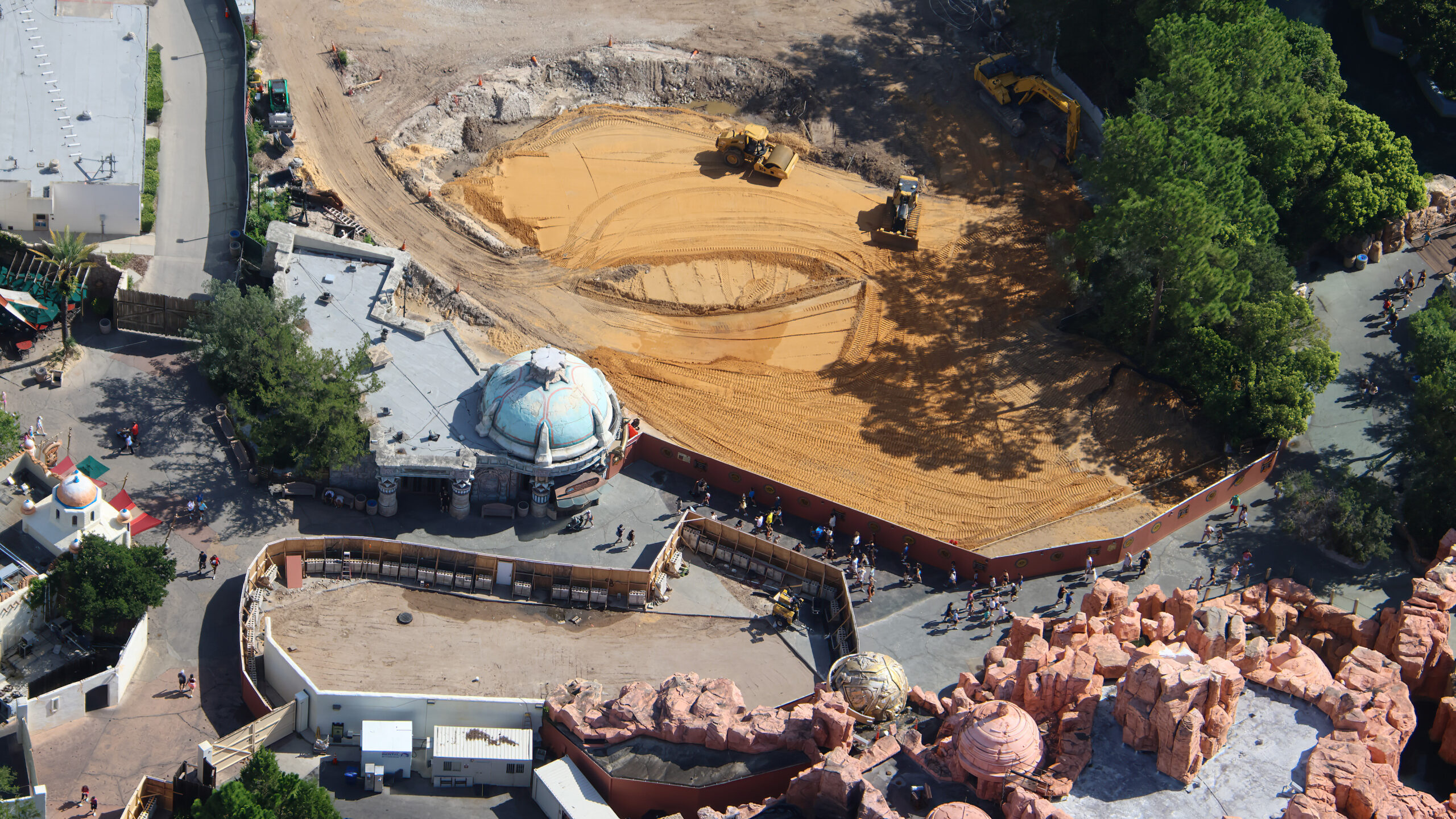 Aerial view of a large kidney shaped pad of freshly graded orange sand with a yellow roller and a dozer working on it, beside a teal domed building, with rust red construction walls curving around and guests walking past
