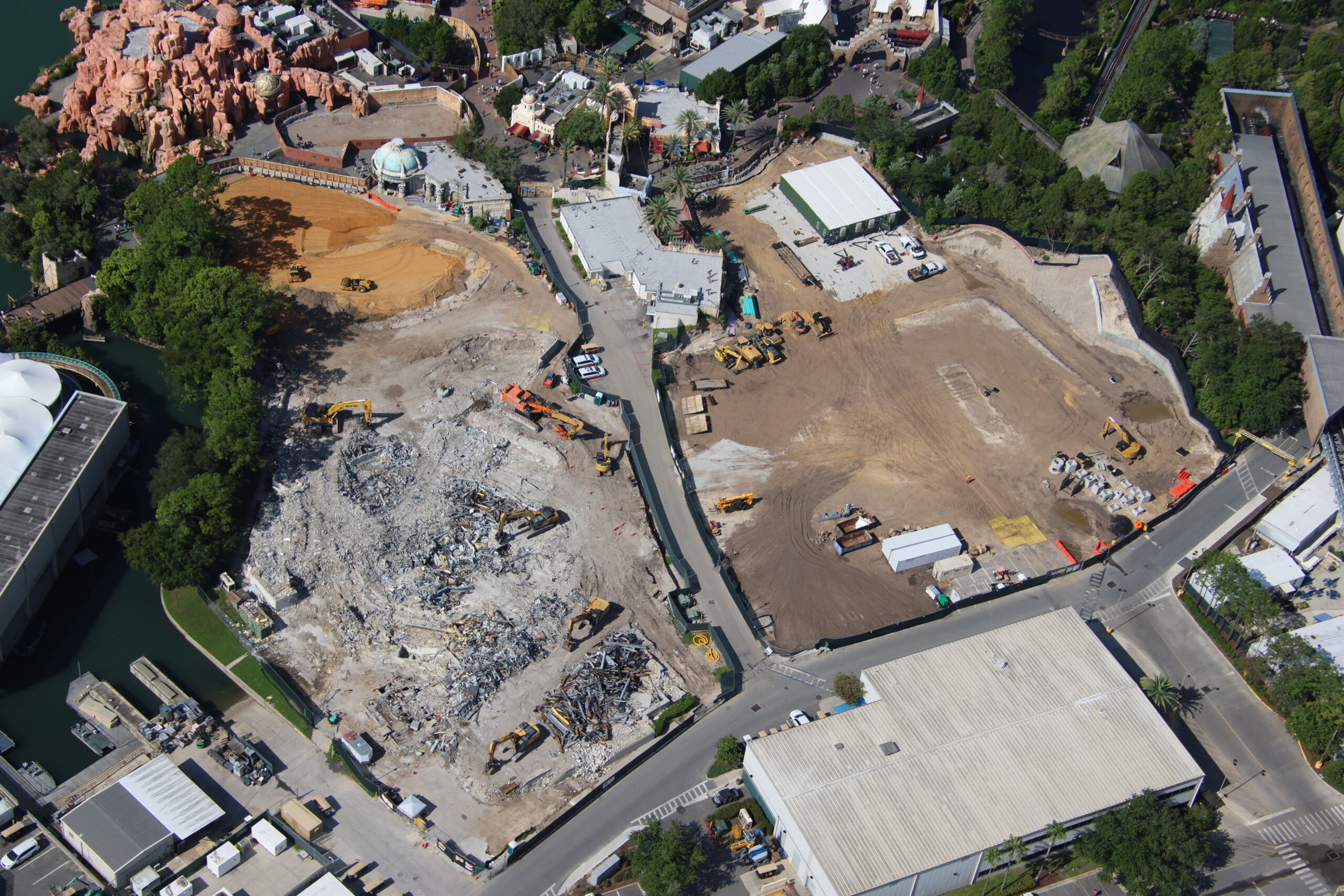 Wide aerial view showing a large grey concrete rubble field with several excavators working in it and a sorted pile of twisted structural steel, beside a large flat bare dirt lot with trailers, a white tent and heavy equipment
