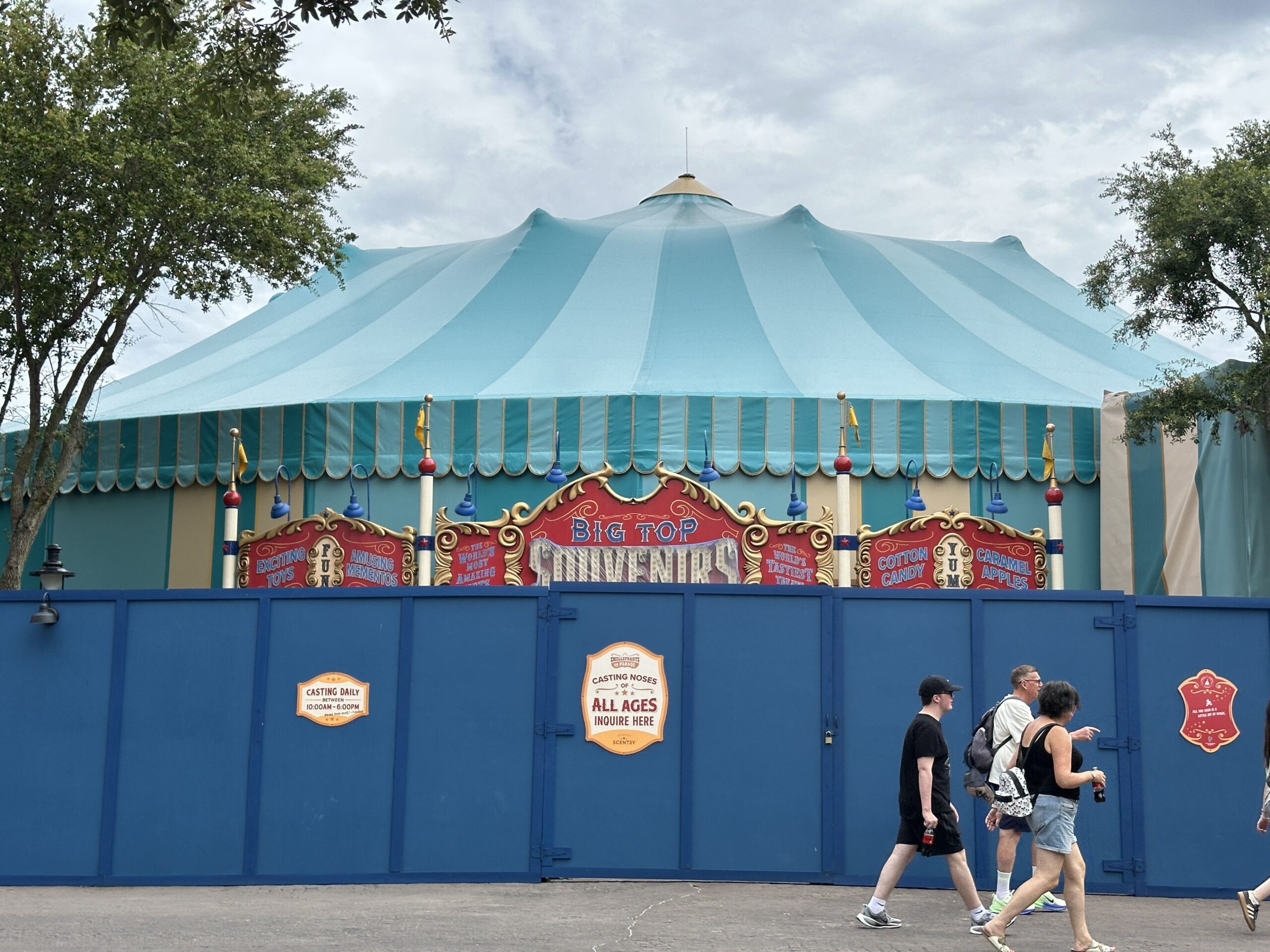 The teal big top tent behind a blue construction wall, with white plastic covering part of the red Big Top Souvenirs marquee sign