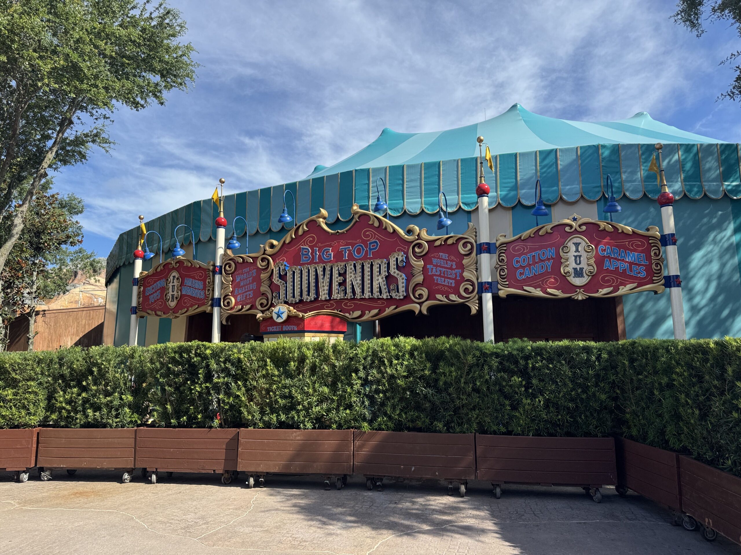 The fully revealed red and gold Big Top Souvenirs marquee with chase light lettering, in front of the teal big top tent, with rolling wooden planters and hedges across the front