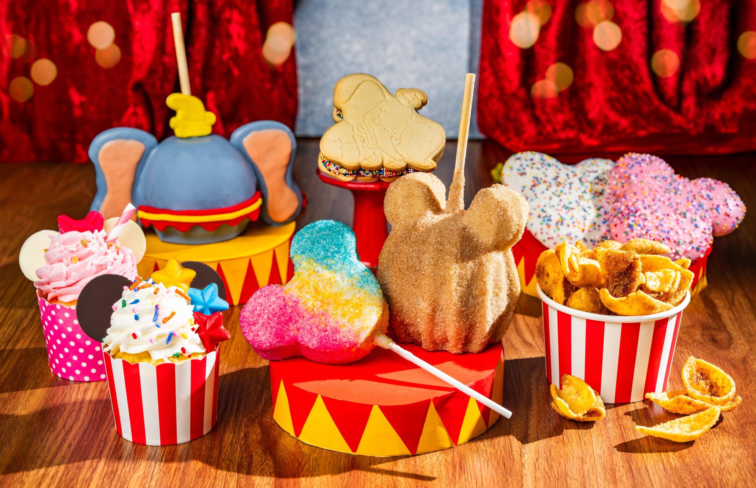 Circus themed Disney treats arranged on a wooden floor in front of a red curtain, including a Dumbo caramel apple, a Mickey shaped churro caramel apple, sprinkle covered marshmallow pops and a striped cup of cinnamon chips